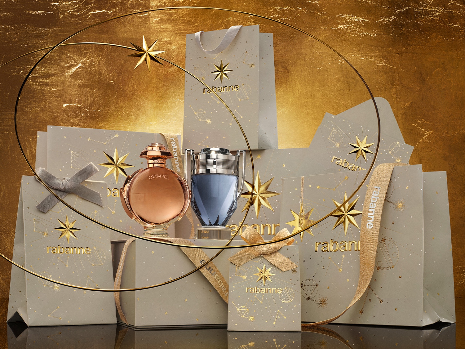 Zlatavé pozadí s dárkovými taškami a krabicemi Rabanne, zdobenými hvězdami a souhvězdími, s parfémy Olympea a Invictus v popředí.