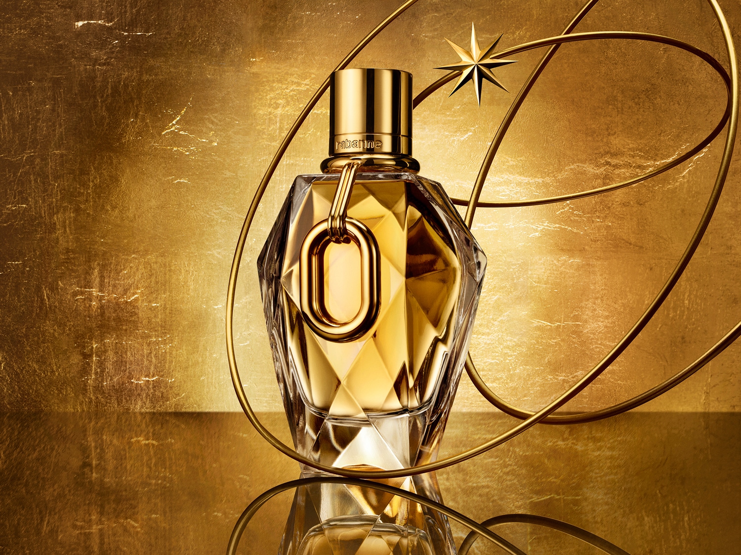 Zlatistý flakón parfému v tvare diamantu s ozdobným zlatým prstencom a hviezdou na pozadí so zlatými líniami.