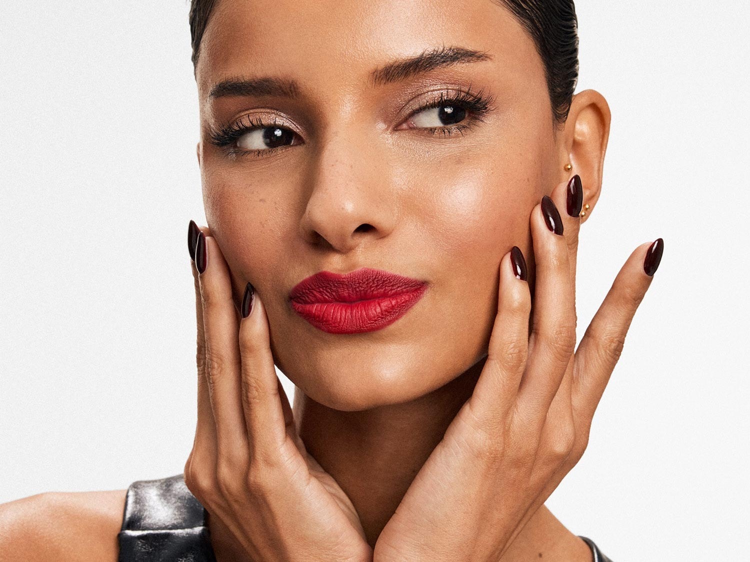 Nahaufnahme einer Frau mit roten Lippen, schimmerndem Lidschatten und dunkelroten Nägeln, die ihr Gesicht berührt und einen eleganten und selbstbewussten Look präsentiert.