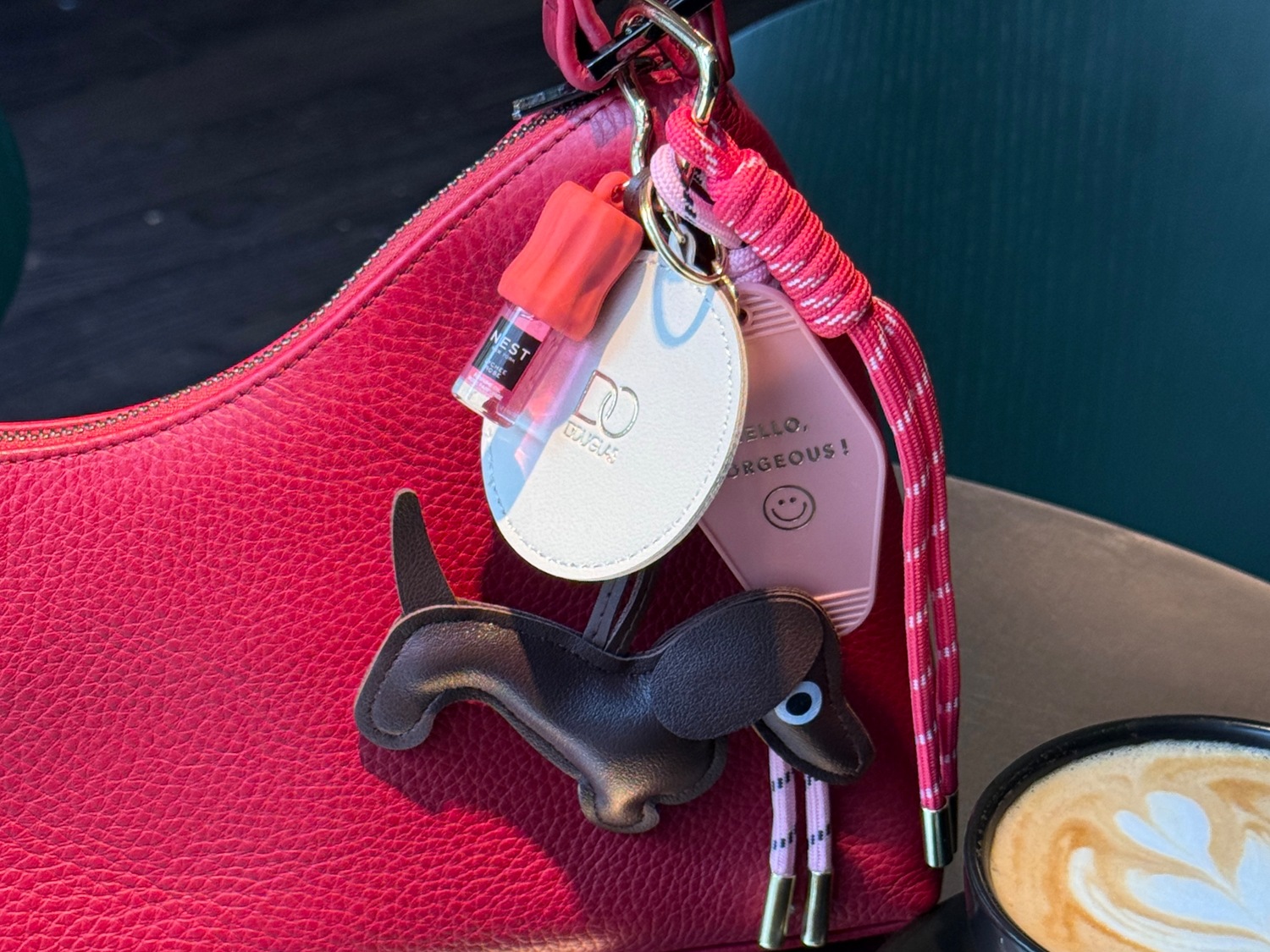 Nahaufnahme einer roten Handtasche mit Anhängern, darunter ein Dackel, ein Nest-Parfüm und ein Douglas-Logo.