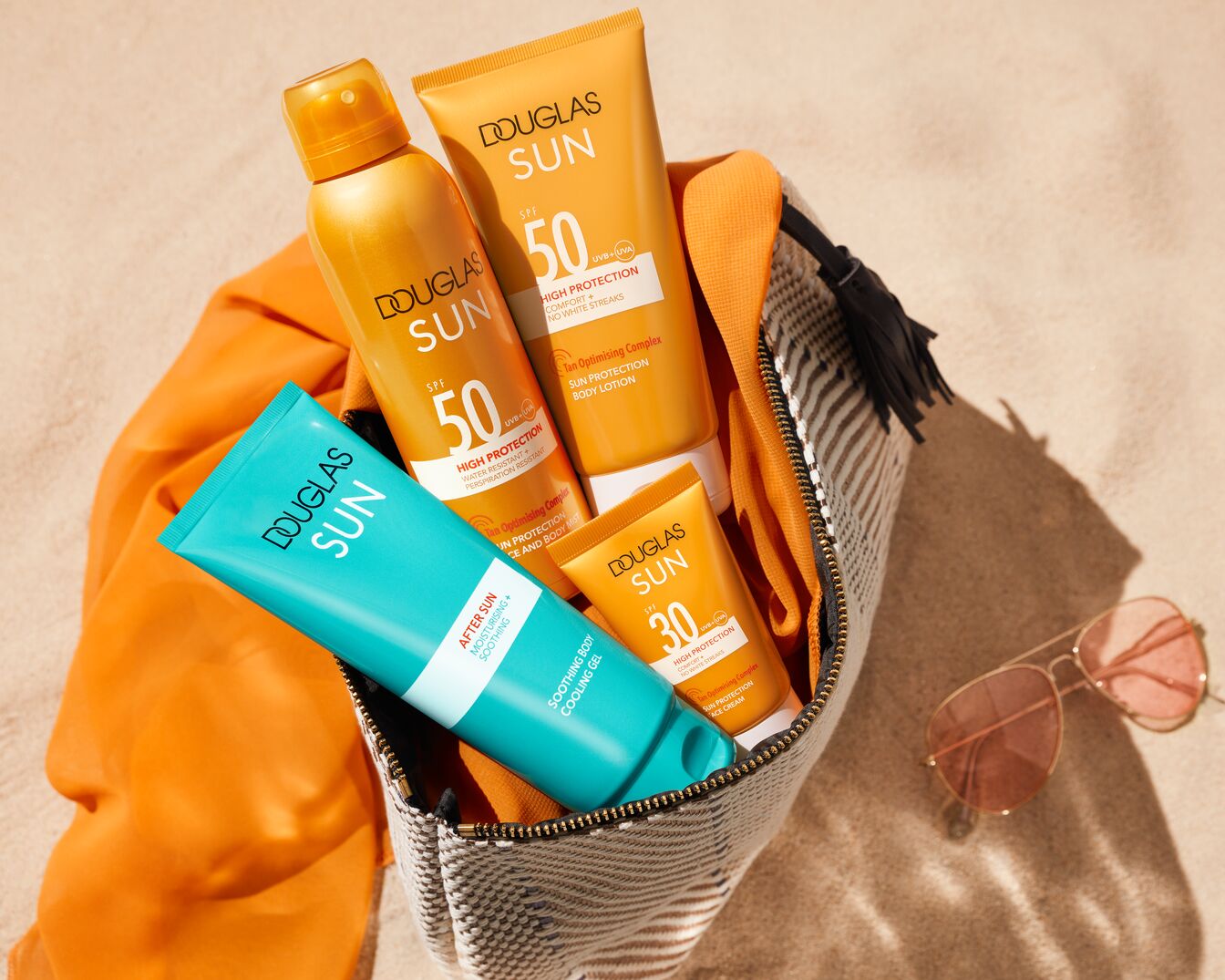 Paplūdimio krepšyje matomi „Douglas Sun“ saulės priežiūros produktai: purškiklis ir losjonas SPF 50, losjonas SPF 30 ir raminamasis gelis po saulės vonių.