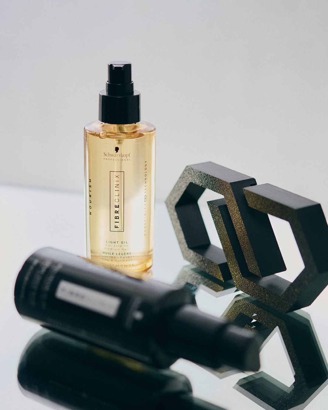Butelka złotego olejku do włosów Schwarzkopf Fibre Clinix Light Oil, przeznaczona do włosów cienkich i normalnych, obok czarnego, geometrycznego elementu dekoracyjnego.
