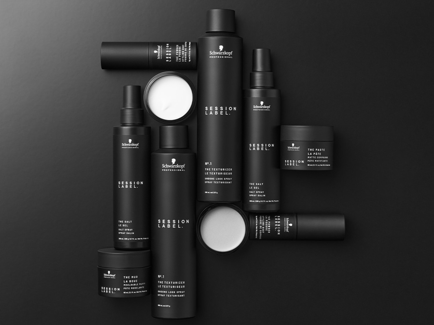 Na czarnym tle leżą czarne opakowania produktów do włosów Schwarzkopf Session Label, w tym spraye, pasty i pudry, tworząc elegancką kompozycję.