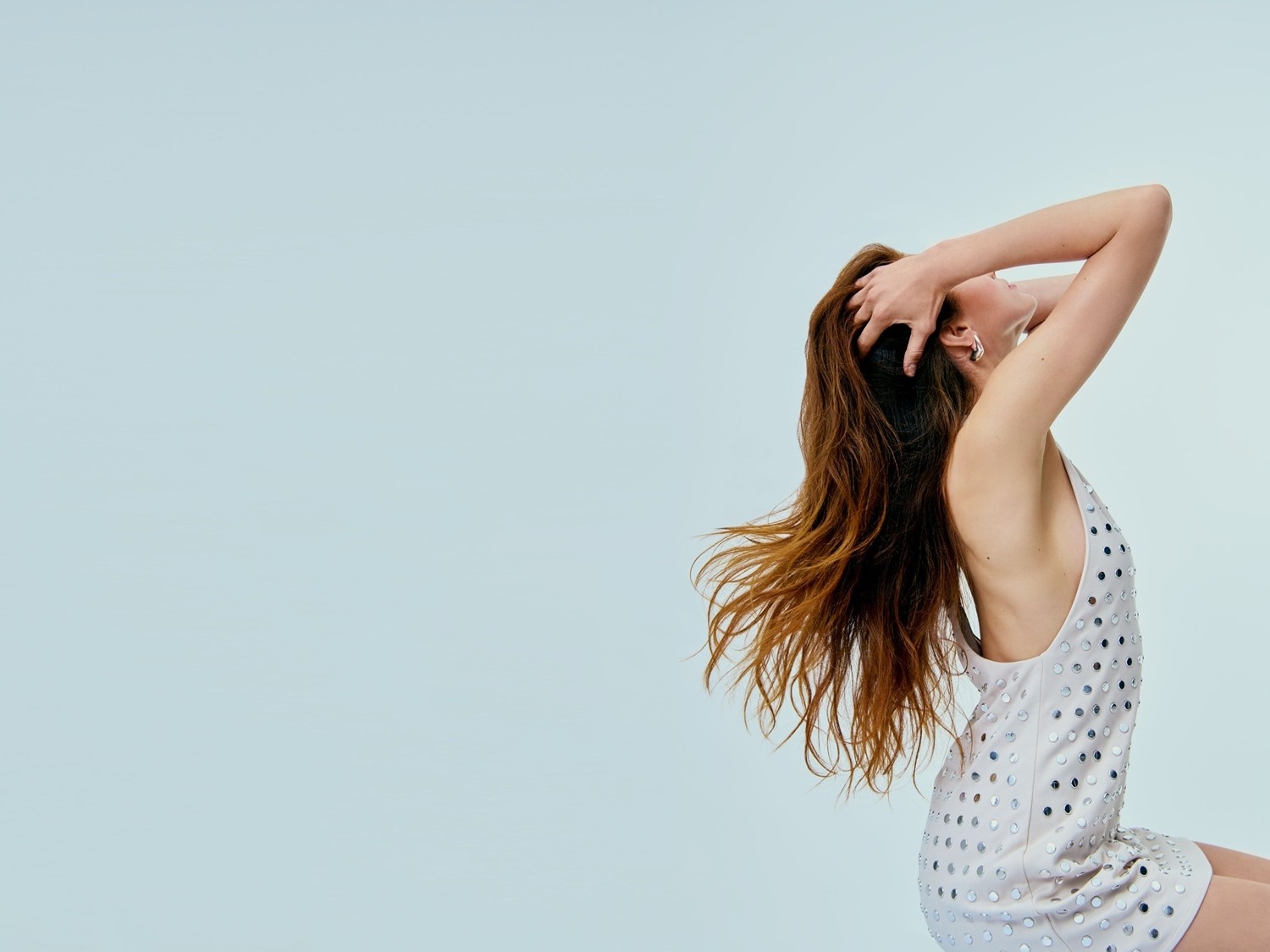 Eine Frau mit langen, gewellten, braunen Haaren, die ein ärmelloses, kurzes, helles Kleid mit silbernen Pailletten trägt, posiert mit erhobenen Armen und nach hinten geneigtem Kopf.