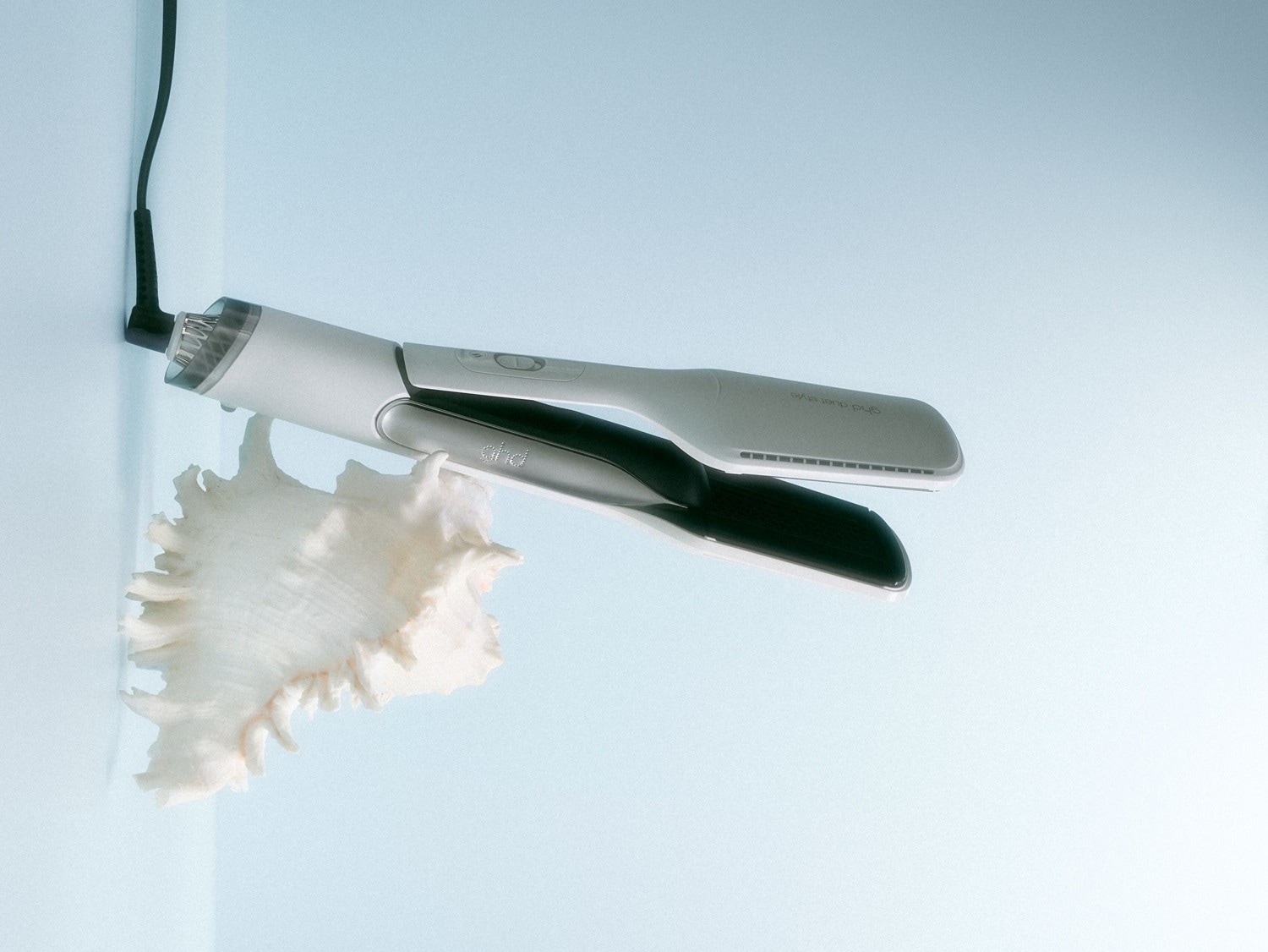 Das Glätteisen in Weiß und Silber von GHD, mit einem schwarzen Kabel und einer großen weißen Muschel, verspricht ein Styling wie am Meer.
