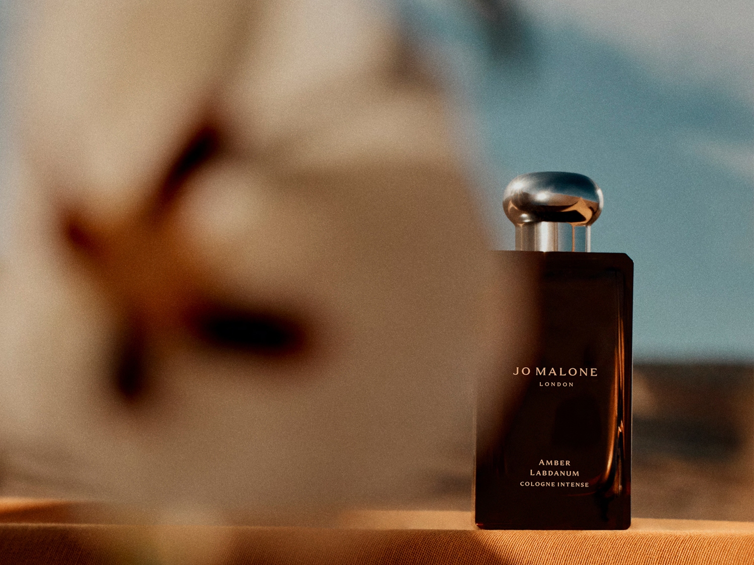 Jo Malone London Amber Labdanum Cologne Intense in einem dunklen, rechteckigen Flakon mit silbernem Verschluss; im Vordergrund ist unscharf eine Blüte zu sehen.