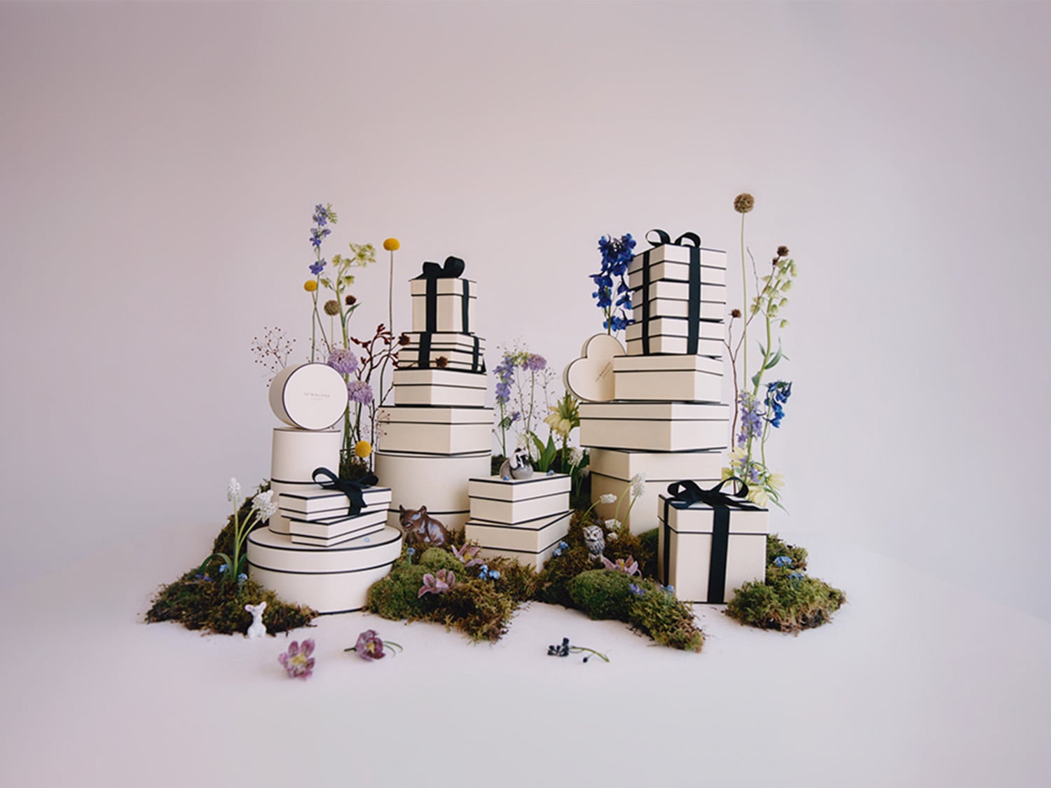 Eine Anordnung von Jo Malone London Geschenkboxen in verschiedenen Formen und Größen, umgeben von Blumen und Moos.