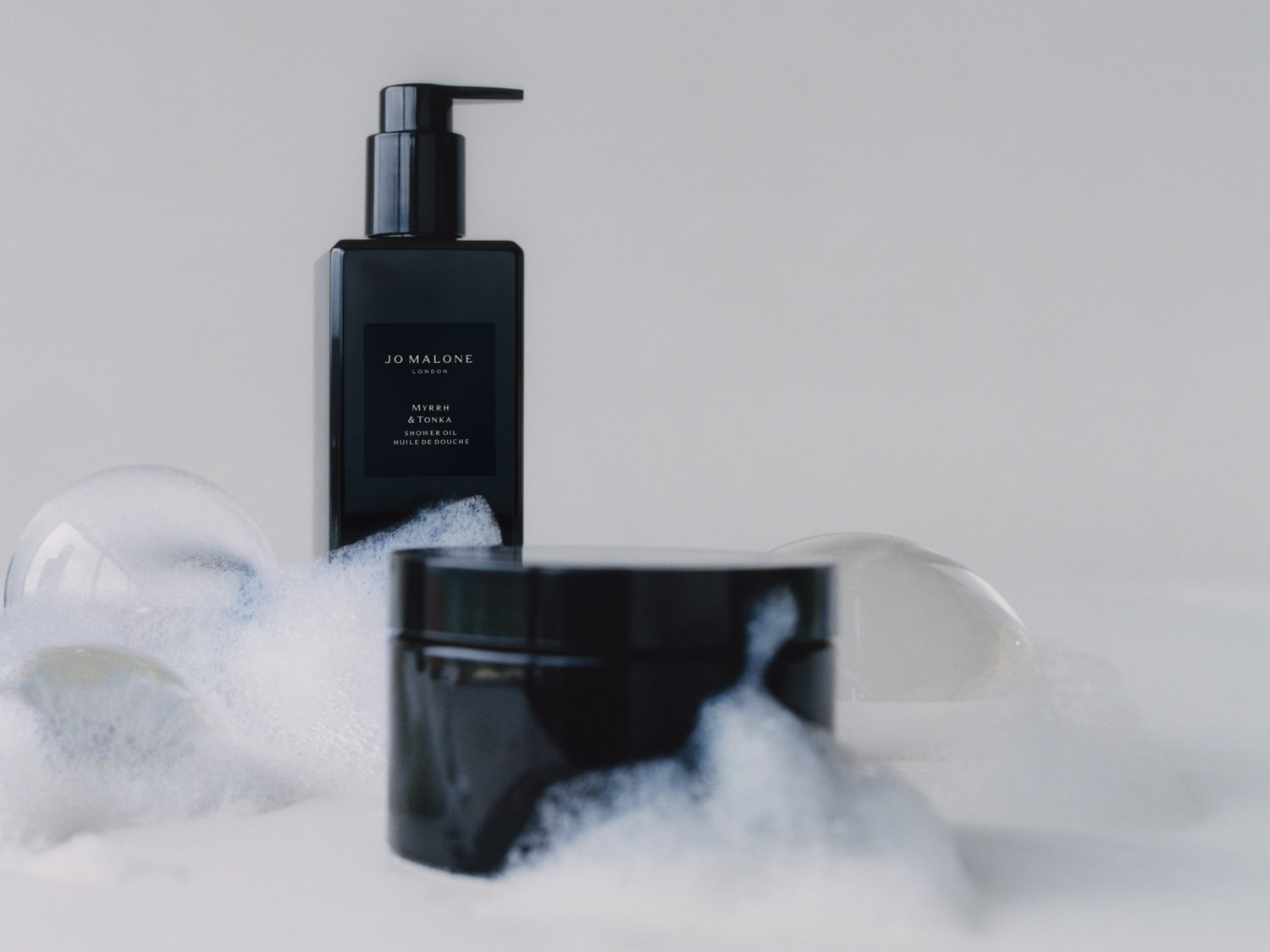 Jo Malone London Myrrh & Tonka Duschöl in einer eleganten, dunklen Flasche mit Pumpspender, umgeben von einer dunklen Dose und Schaum.