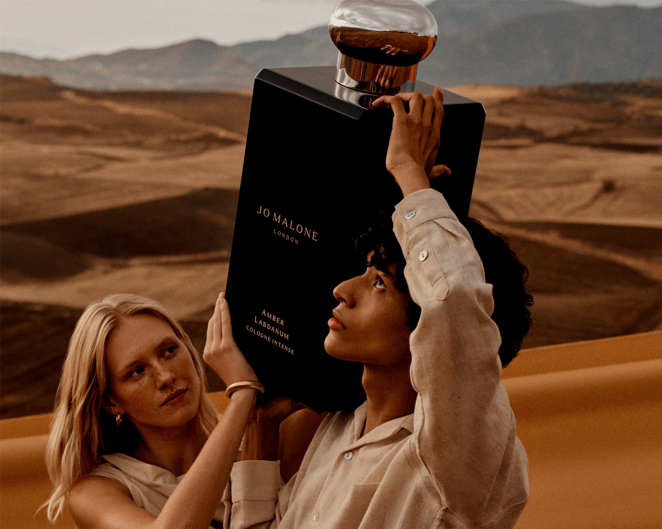 Ein riesiger, schwarzer Flakon von Jo Malone Amber Labdanum Cologne Intense wird von zwei Personen in einer sandigen Wüstenlandschaft gehalten.