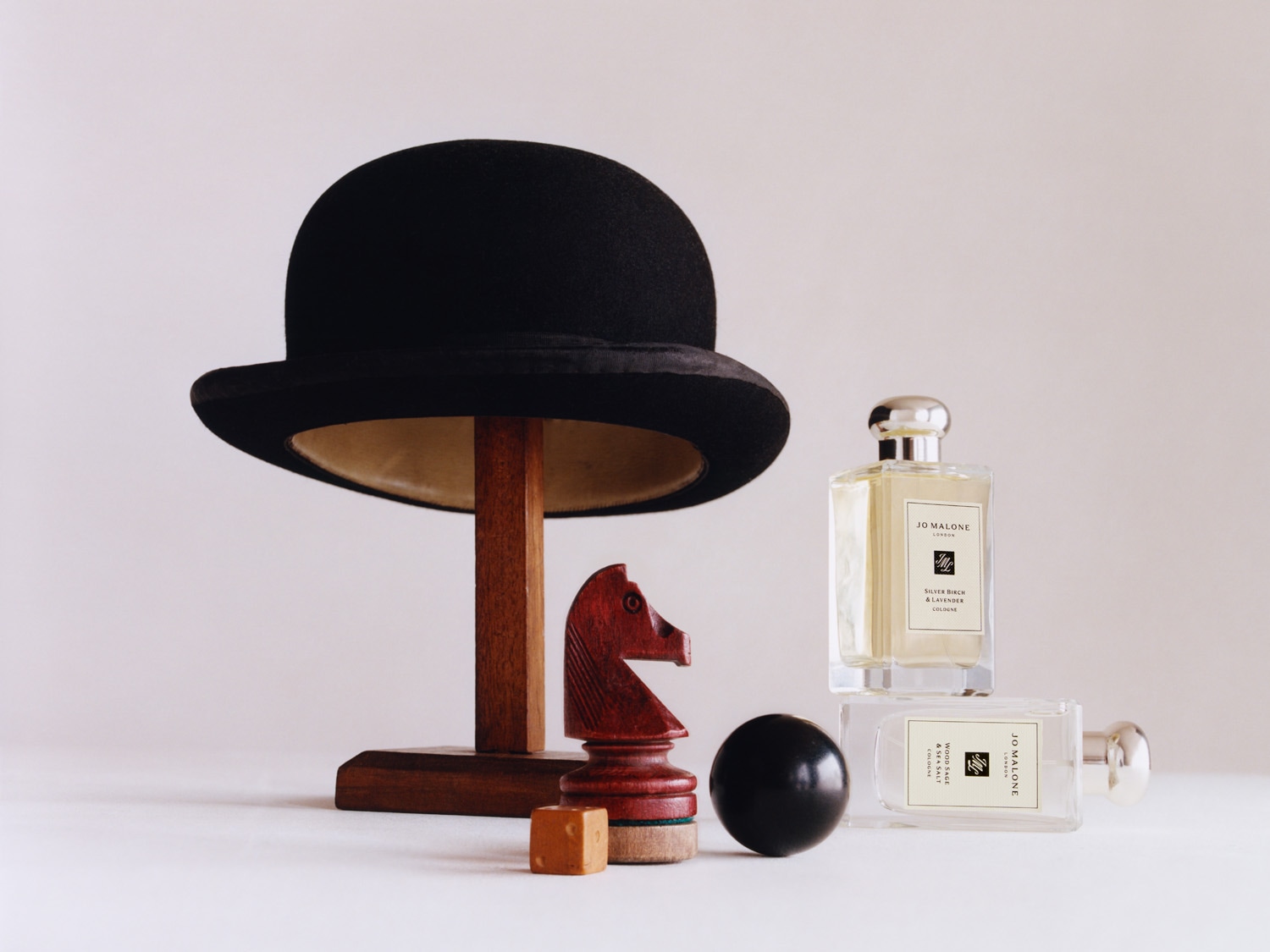 Ein Arrangement mit einem schwarzen Hut auf einem Holzständer, einer Schachfigur, einem Würfel, einer schwarzen Kugel und zwei Flakons Jo Malone Düfte.