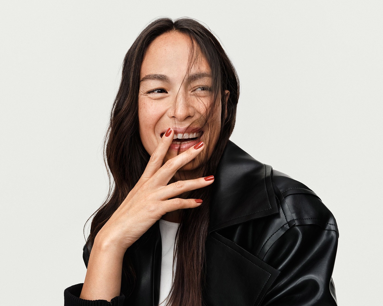 Une femme souriante, les doigts vernis d'un rouge éclatant près de la bouche, vêtue d'une veste en cuir noire élégante.