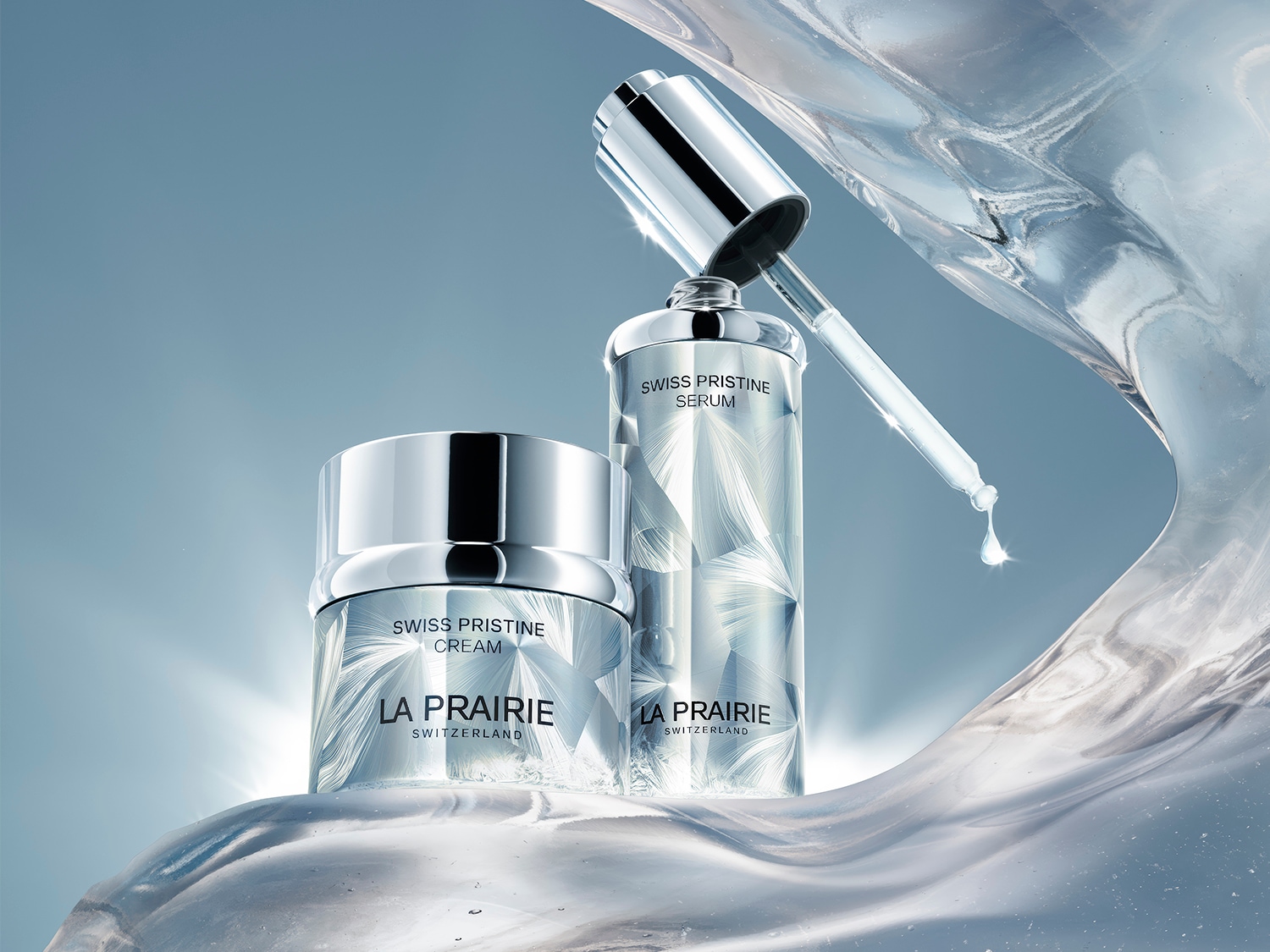 La Prairie Swiss Pristine Cream i Serum, w eleganckich srebrnych opakowaniach z białymi wzorami, stoją na błyszczącej, falującej powierzchni, a z pipety serum spływa kropla.