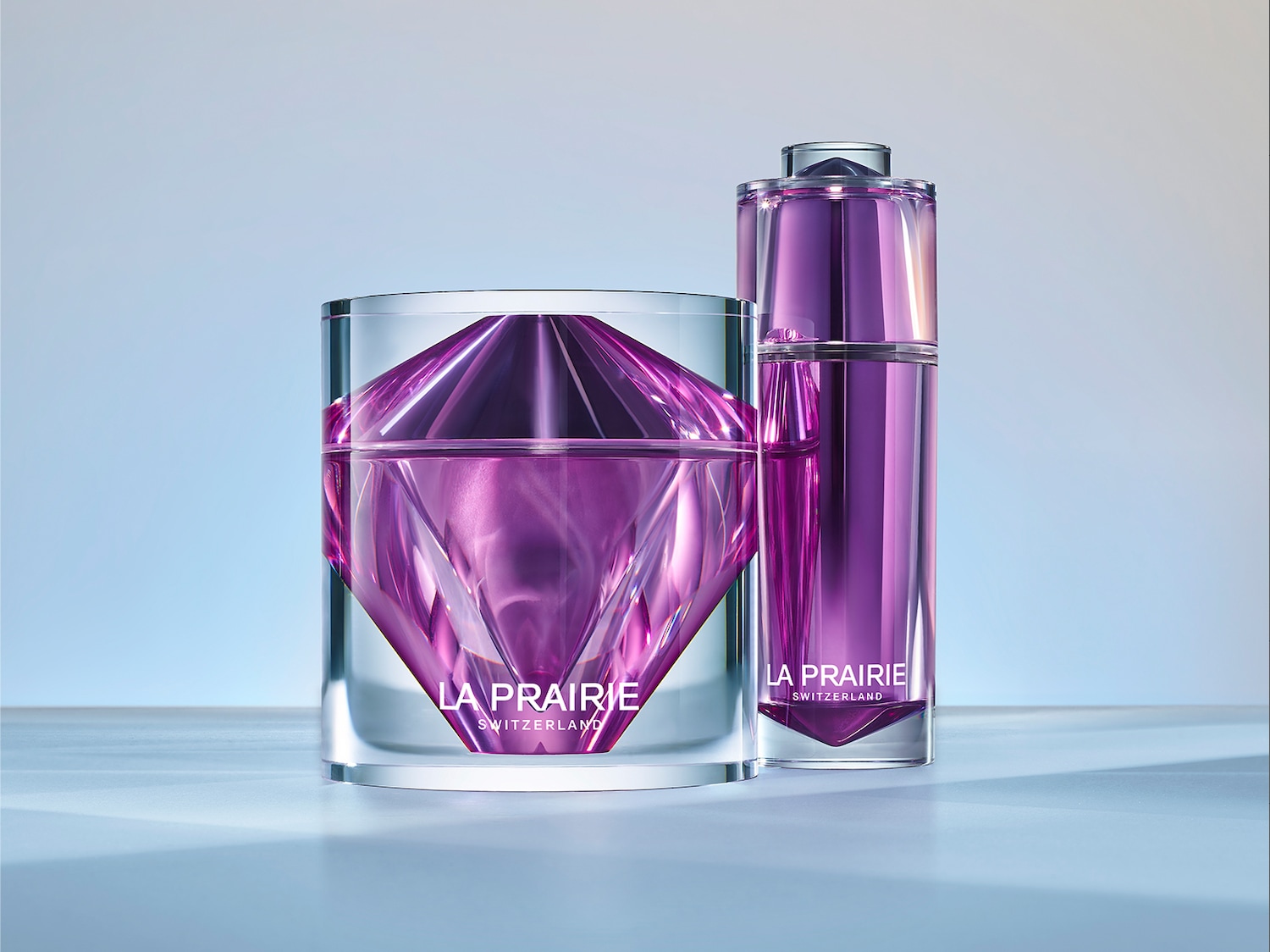 Dwa produkty La Prairie w przezroczystych opakowaniach z fioletową zawartością, krem w słoiczku w kształcie diamentu i serum w butelce z dozownikiem.