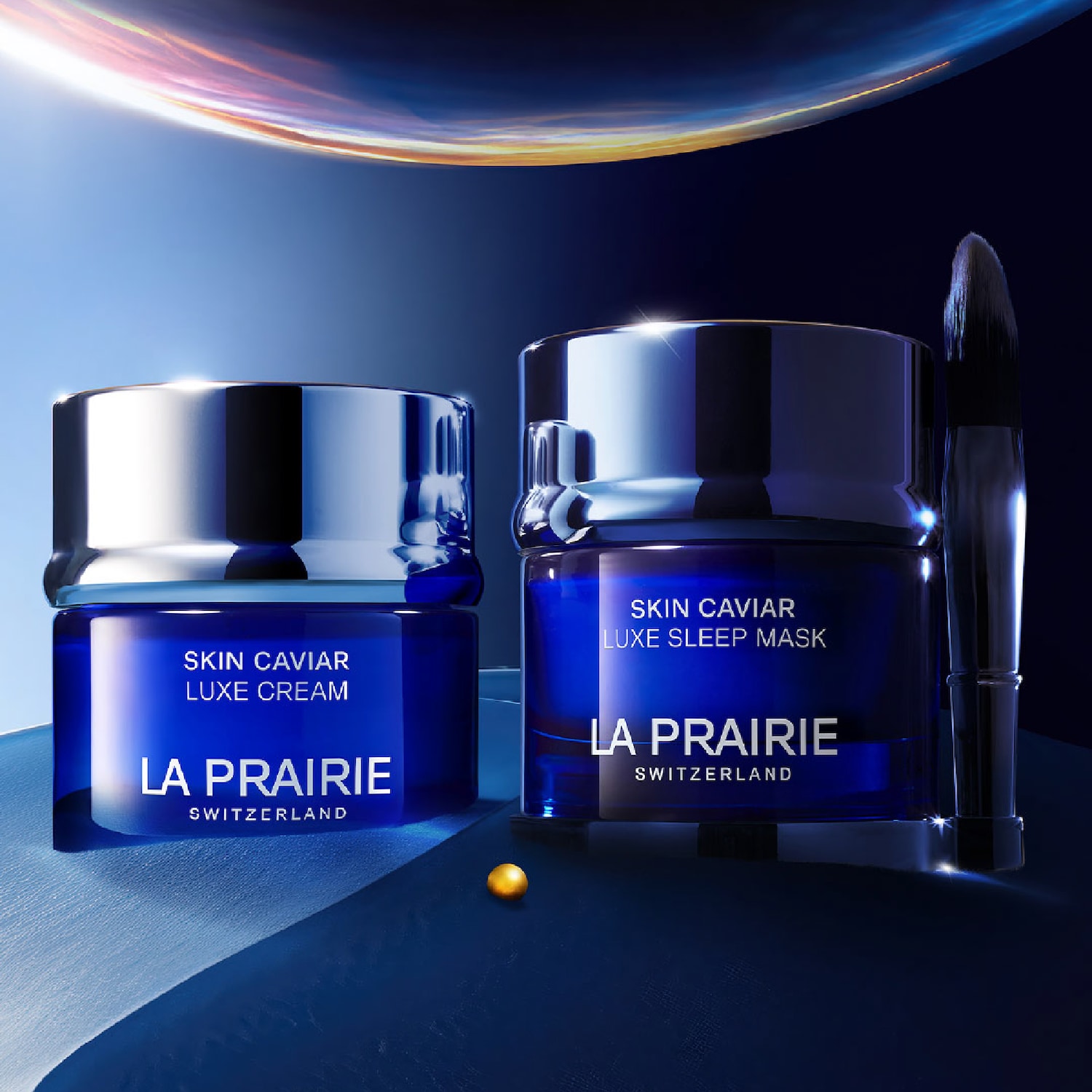 Dwa niebieskie słoiczki La Prairie Skin Caviar Luxe Cream i Luxe Sleep Mask, obok pędzelek do nakładania maseczki, na tle nocnego nieba.