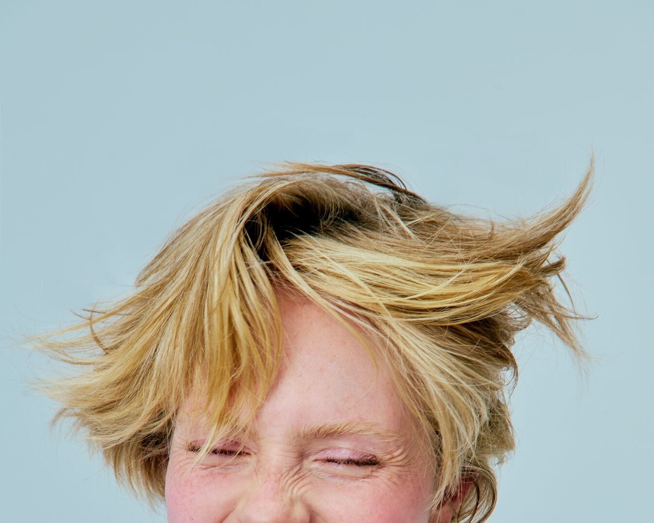Nahaufnahme einer Frau mit blonden, zerzausten Haaren und zusammengekniffenen Augen vor hellblauem Hintergrund.