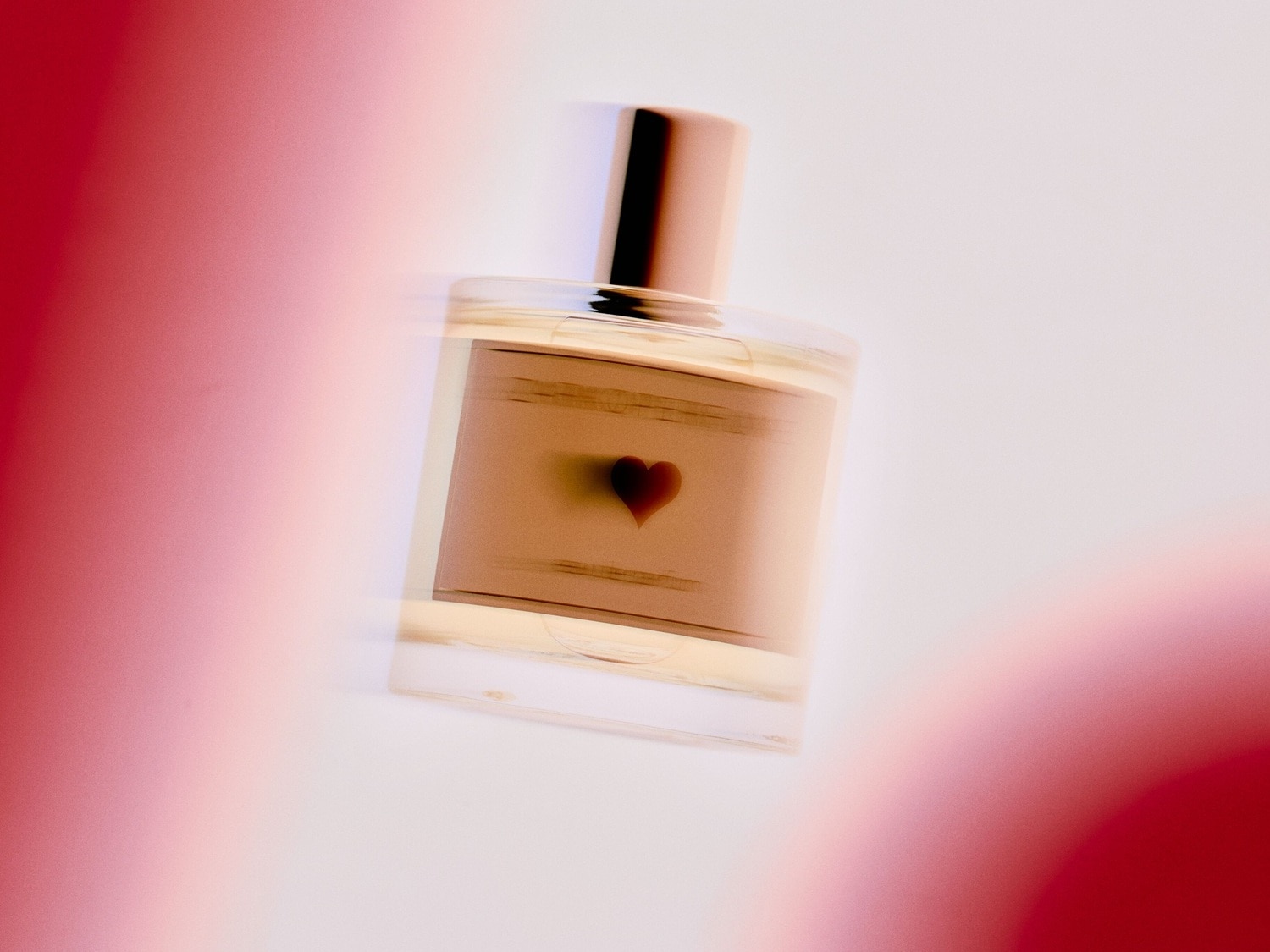 Ein rechteckiger, transparenter Parfümflakon mit einem hellbraunen Etikett, das ein kleines Herz zeigt, und einem bronzefarbenen Verschluss.