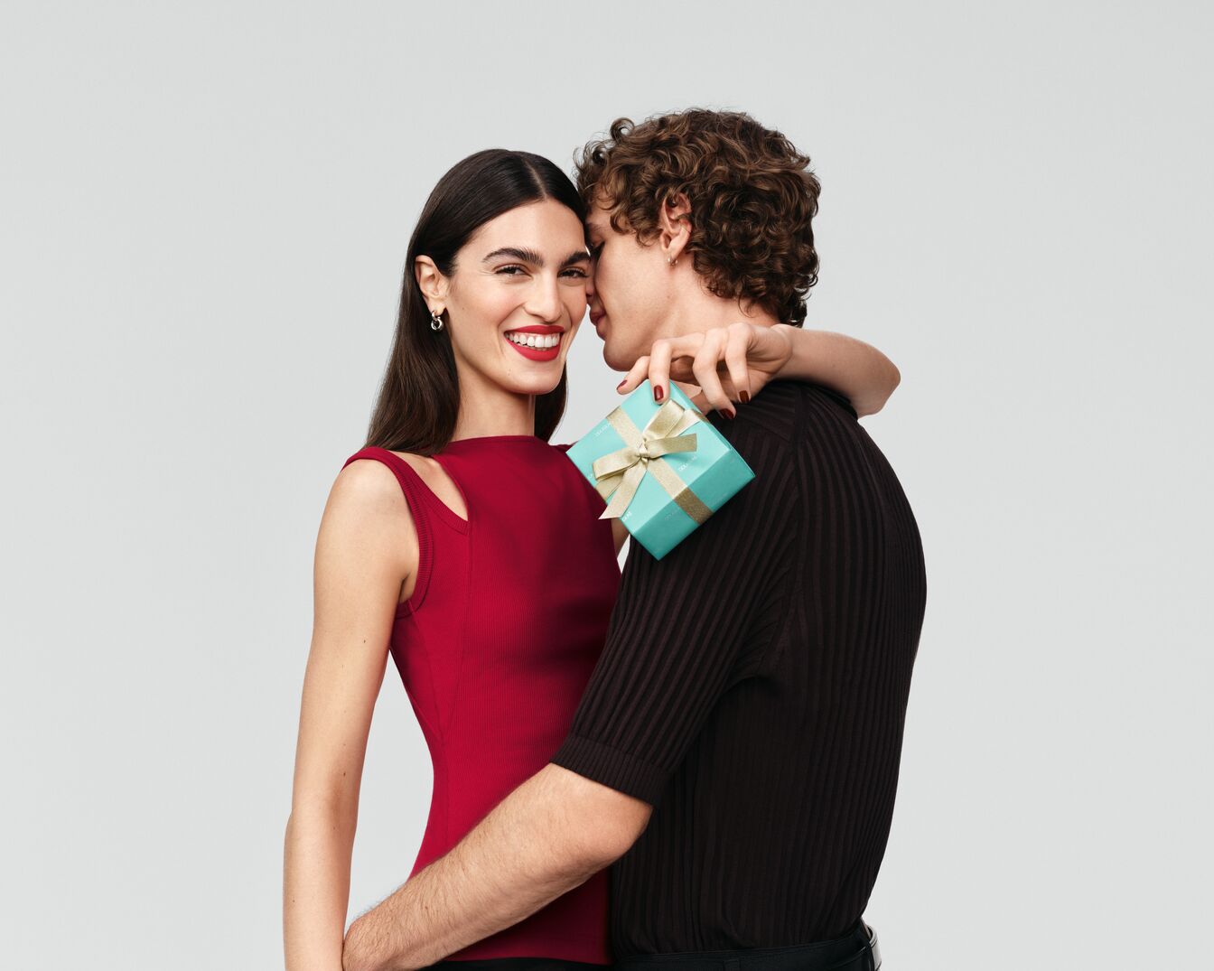 Ein Paar umarmt sich, wobei die Frau ein strahlendes Lächeln und ein rotes Oberteil trägt, während der Mann eine kleine, hellblaue Geschenkbox mit goldener Schleife hält.