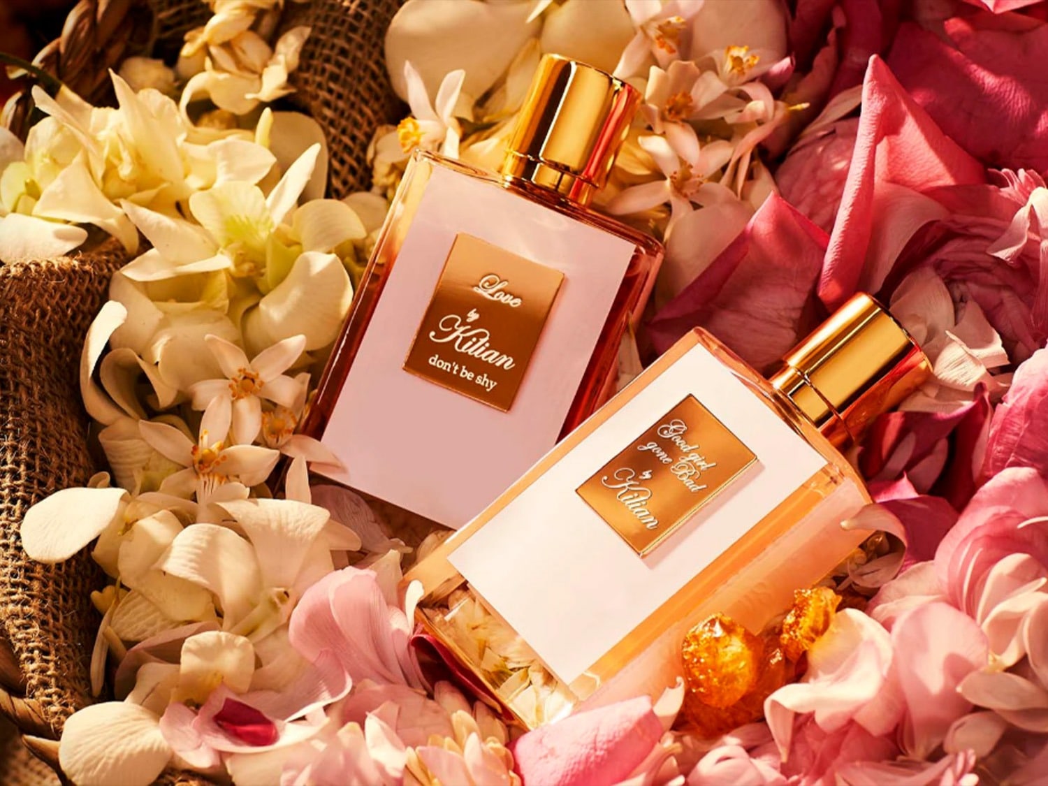 Deux parfums Kilian, l'un rose pâle avec l'inscription « Love, don't be shy », l'autre pêche avec « Good girl gone Bad », reposent sur un lit de pétales de fleurs blanches et roses.