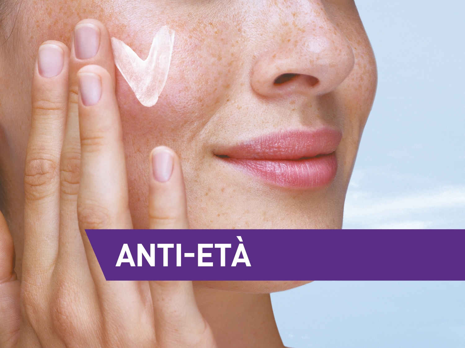 Primo piano di una donna che applica una crema bianca sul viso, con la scritta 'ANTI-ETÀ' in viola.