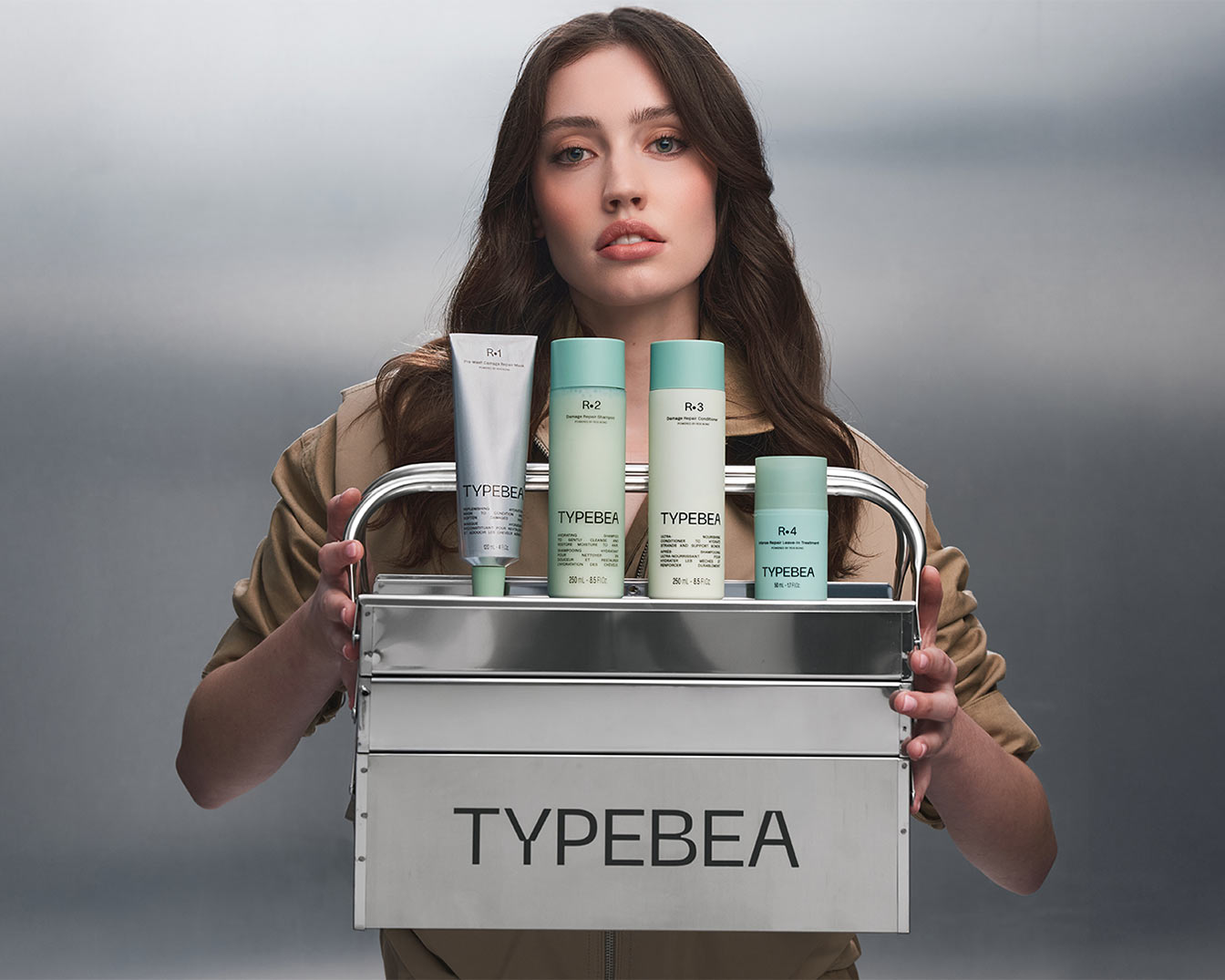 Eine Frau hält einen silbernen Werkzeugkasten mit vier TYPEBEA-Haarpflegeprodukten, darunter eine Tube und Flaschen in Weiß und Grün.