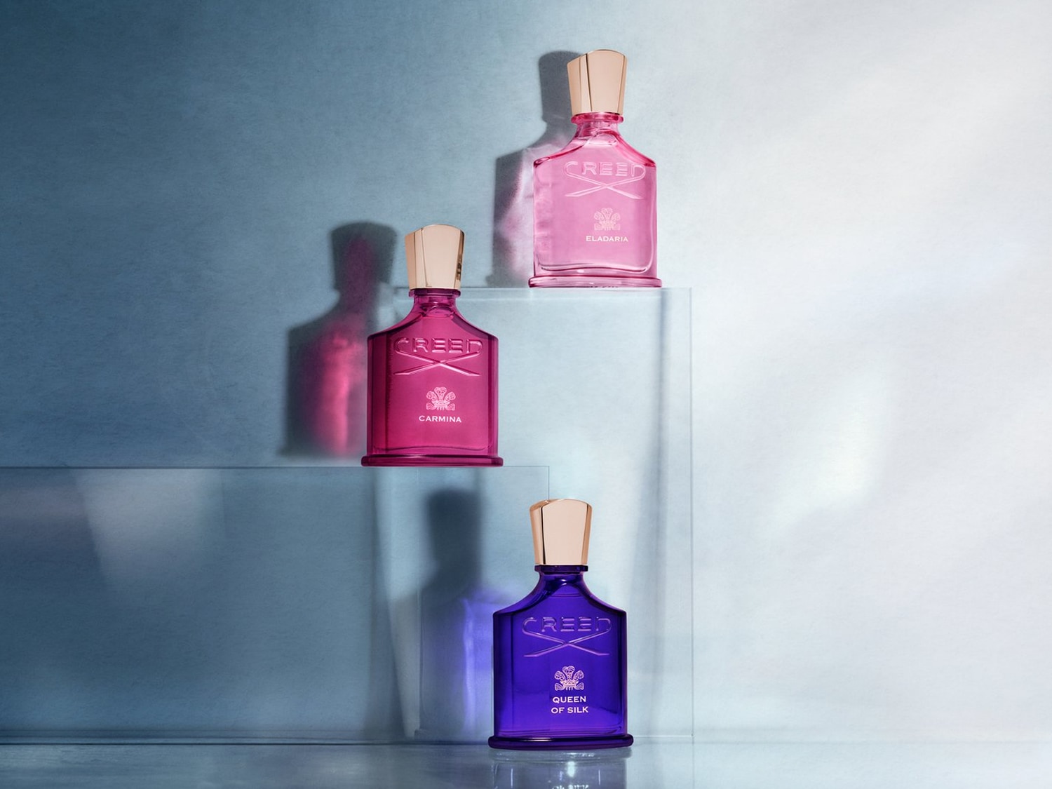 Trzy flakony perfum Creed w odcieniach różu, fuksji i fioletu, każdy z nazwą i złotą nakrętką, ustawione na przezroczystych podstawkach na tle w kolorze lodowego błękitu.