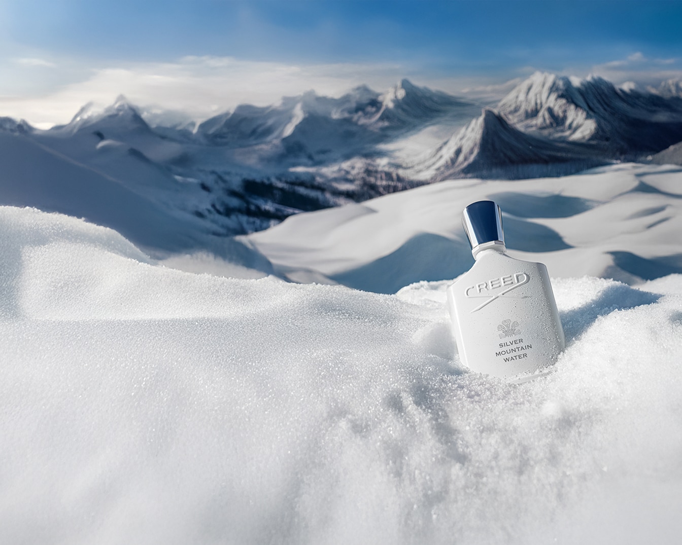Biały flakon perfum Creed Silver Mountain Water stoi na śniegu z ośnieżonymi górami w tle, emanując świeżością i czystością.