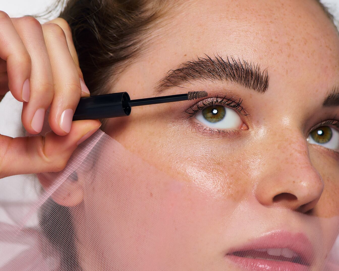 Detailný záber na ženu, ktorá si nanáša hnedú maskaru na obočie pomocou čiernej kefky, s dôrazom na jej zelené oči a pehy.