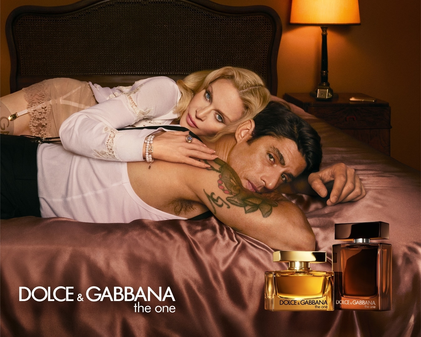 Na reklame Dolce & Gabbana The One leží na posteli pár, v popredí sú dva flakóny vône, jeden zlatý a jeden hnedý.