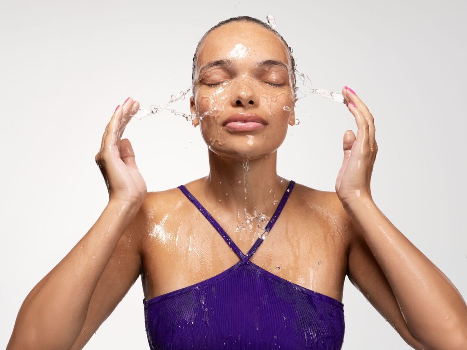 Une femme aux cheveux mouillés et à la peau éclatante se rafraîchit le visage avec de l'eau, portant un haut violet.