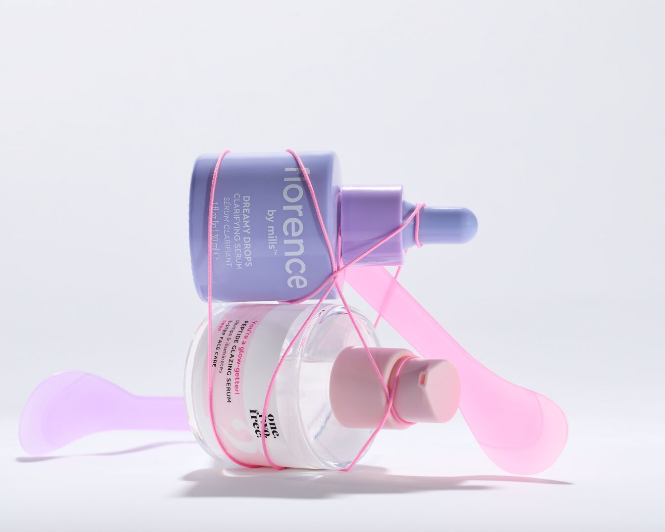 Deux sérums pour le visage, l'un dans un flacon violet "Florence by Mills Dreamy Drops Clarifying Serum", l'autre dans un flacon transparent "One. Two. Free. Peptide Glazing Serum", liés par un ruban rose.