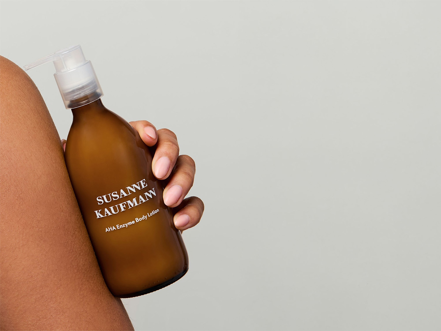 Eine Hand hält eine braune Flasche Susanne Kaufmann AHA Enzym Körperlotion mit einem durchsichtigen Pumpspender.