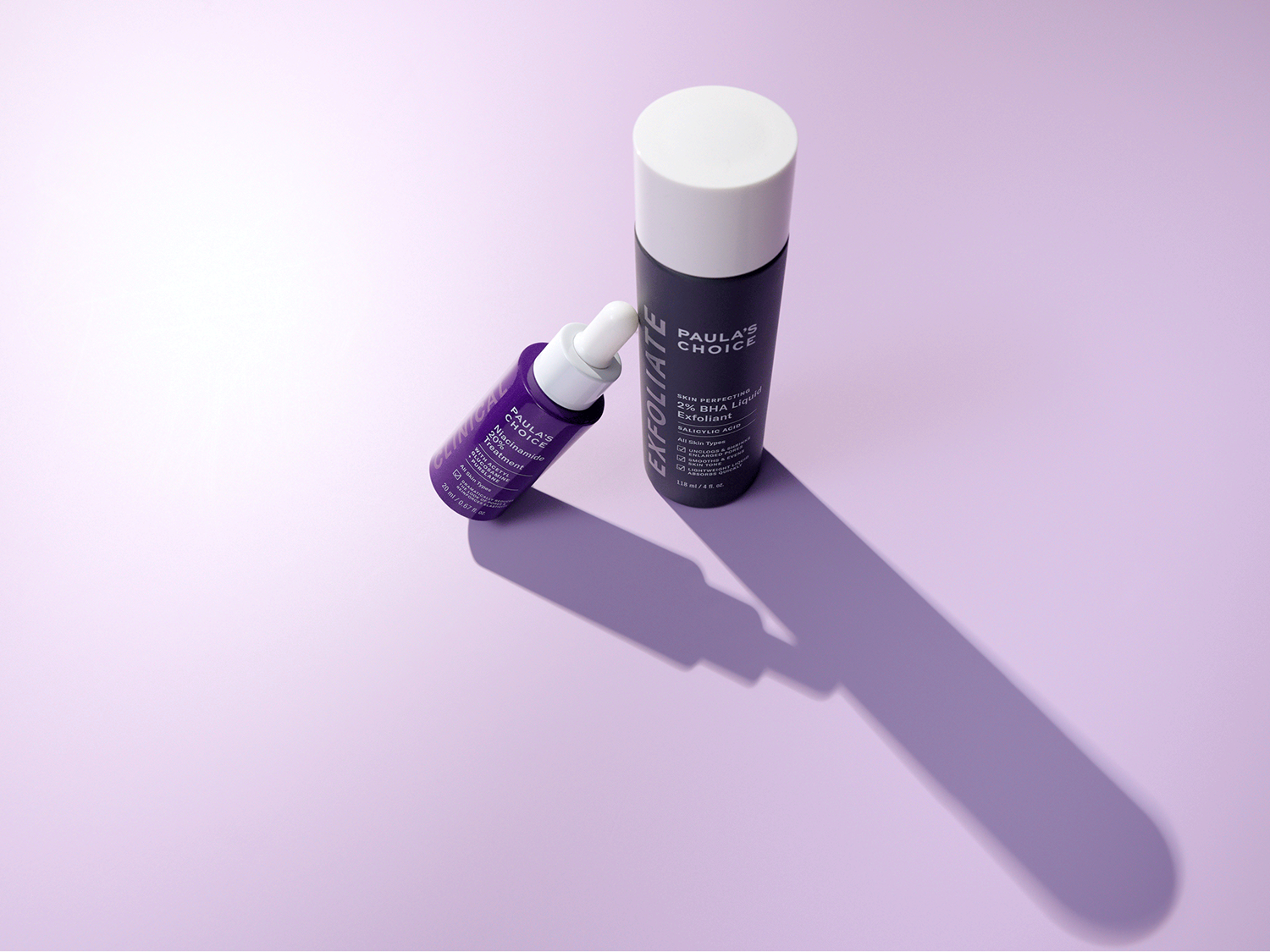Voici le soin Niacinamide 20 % de Paula's Choice dans un flacon violet foncé avec un compte-gouttes blanc, à côté de l'exfoliant liquide 2 % BHA dans un flacon gris foncé avec un bouchon blanc.