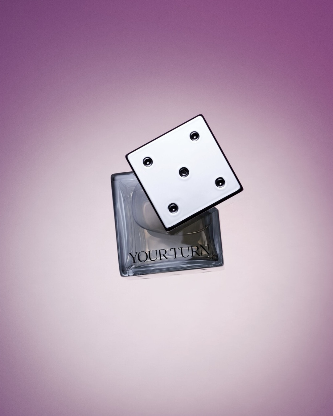 De heldere glazen fles 'YOUR TURN' heeft een dop in de vorm van een witte dobbelsteen met zwarte stippen, tegen een zachtpaarse achtergrond.