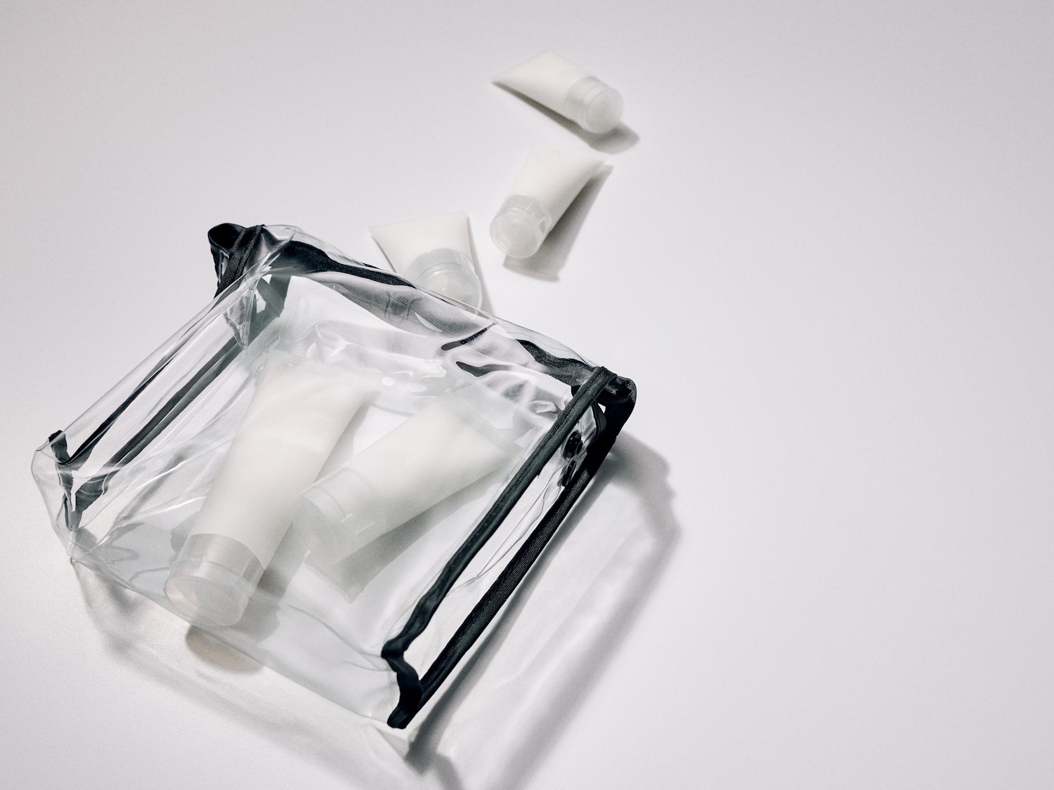 Průhledná kosmetická taška s černým zipem obsahuje několik bílých cestovních tub s kosmetikou.