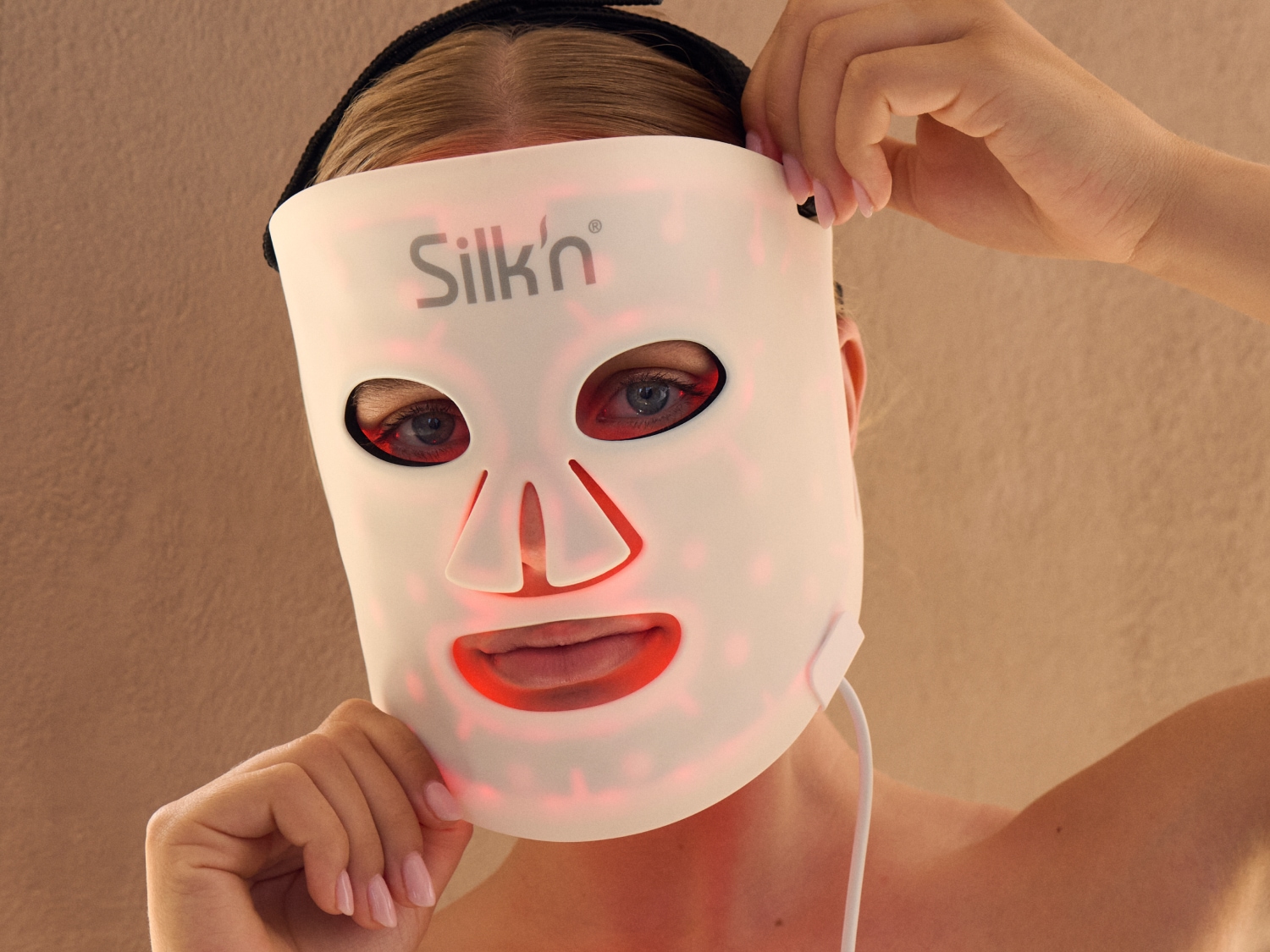Een vrouw houdt een wit Silk'n gezichtsmasker met rode LED-verlichting voor haar gezicht, wat een futuristische en huidverzorgende uitstraling geeft.
