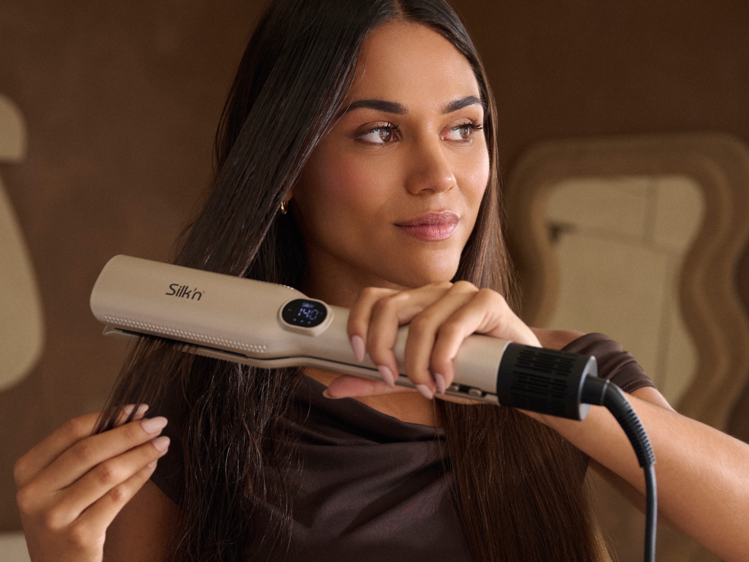 Een vrouw met lang bruin haar gebruikt een Silk'n stijltang met een digitaal display dat 140 graden aangeeft, om haar haar glad te maken.
