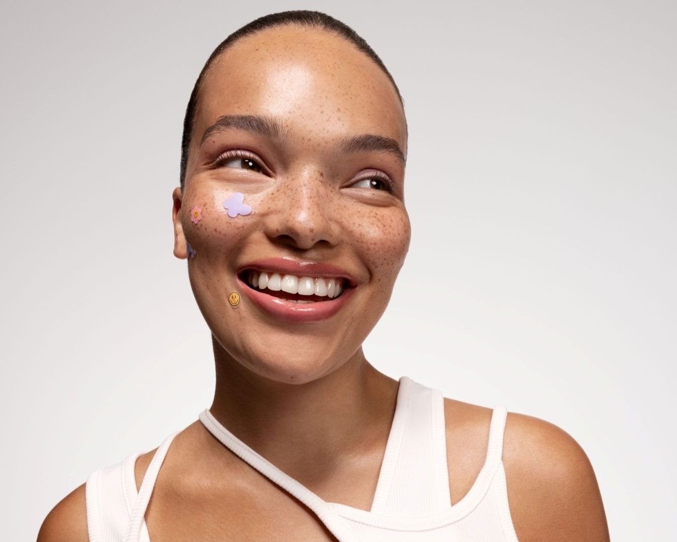 Primo piano di una donna sorridente con lentiggini e adesivi a forma di fiore e faccina sorridente sul viso, che indossa una canotta bianca.