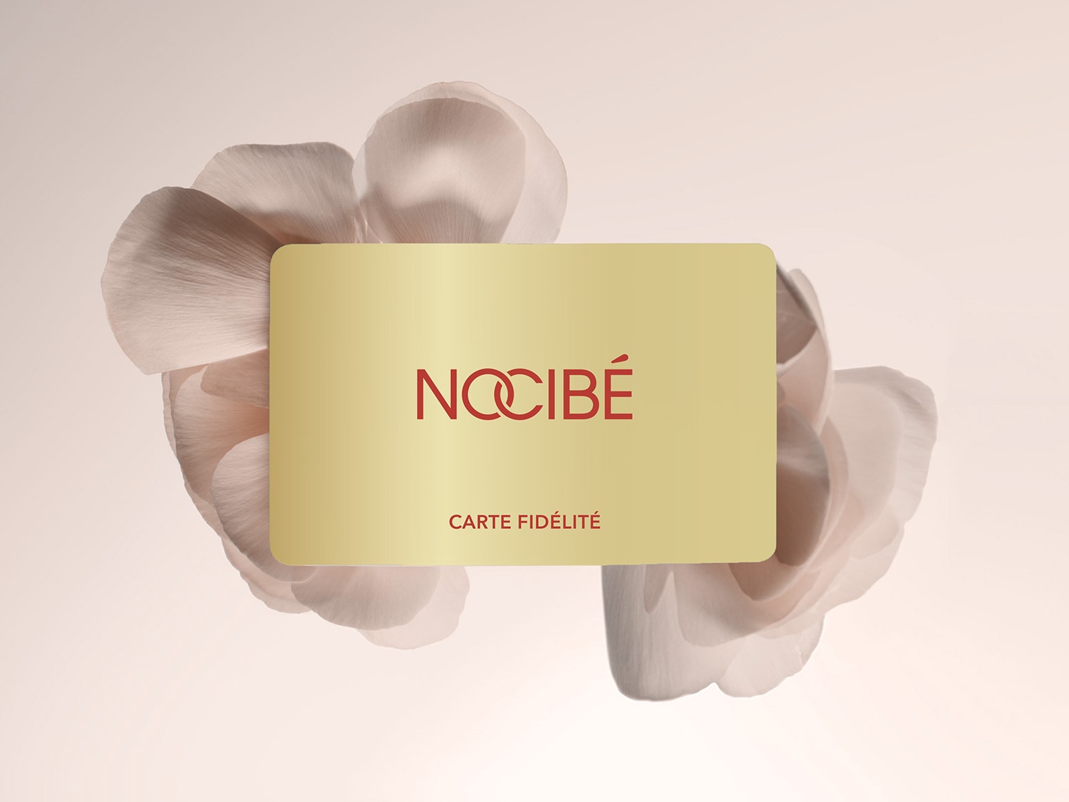 La carte de fidélité Nocibé dorée avec le logo rouge de la marque et le texte "CARTE FIDÉLITÉ" est présentée sur un fond clair orné de pétales de fleurs roses et blanches.