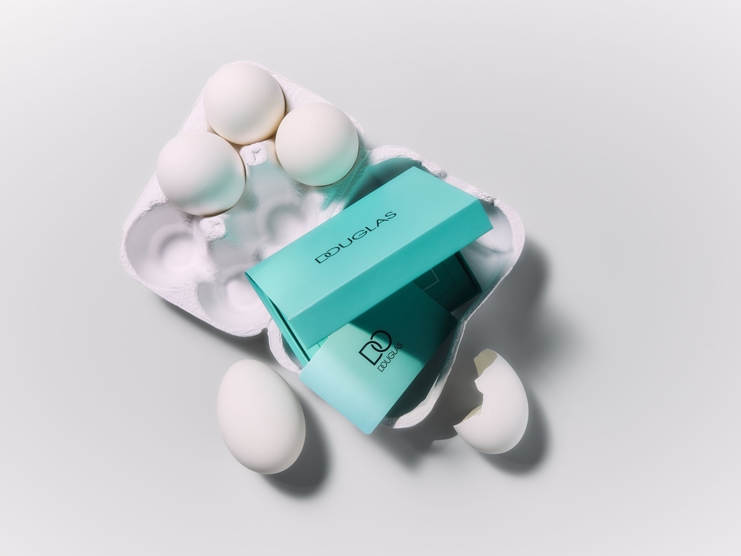 Šviesiame fone pavaizduota kiaušinių dėžutė su keliais baltais kiaušiniais ir firminė turkio spalvos „Douglas“ dovanų dėžutė.