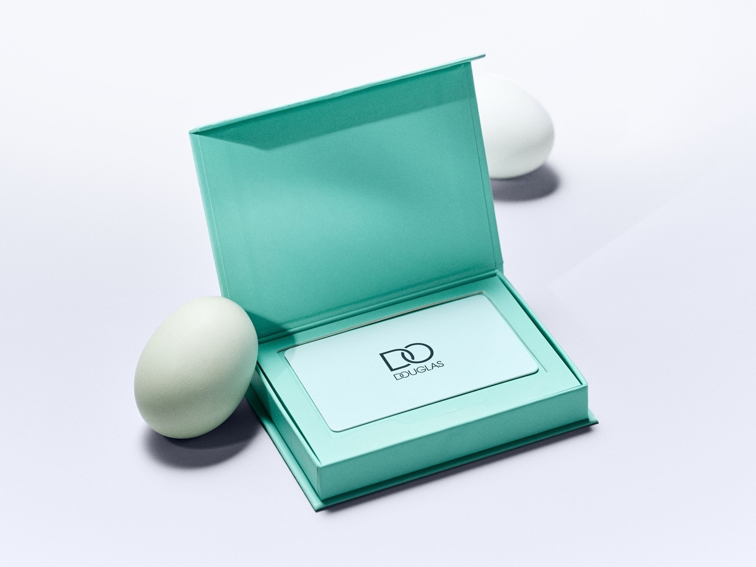 Mėlynos spalvos „Douglas“ dovanų kortelė atvirame dėkle šalia dviejų baltų kiaušinių.