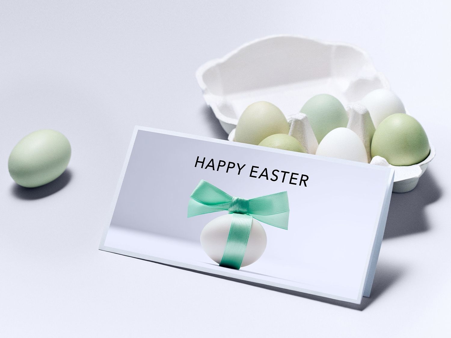 Atvirukas su užrašu „Linksmų Velykų“, papuoštas kiaušiniu su žaliu kaspinu, šalia dėžutė su margučiais – puikus velykinis akcentas.