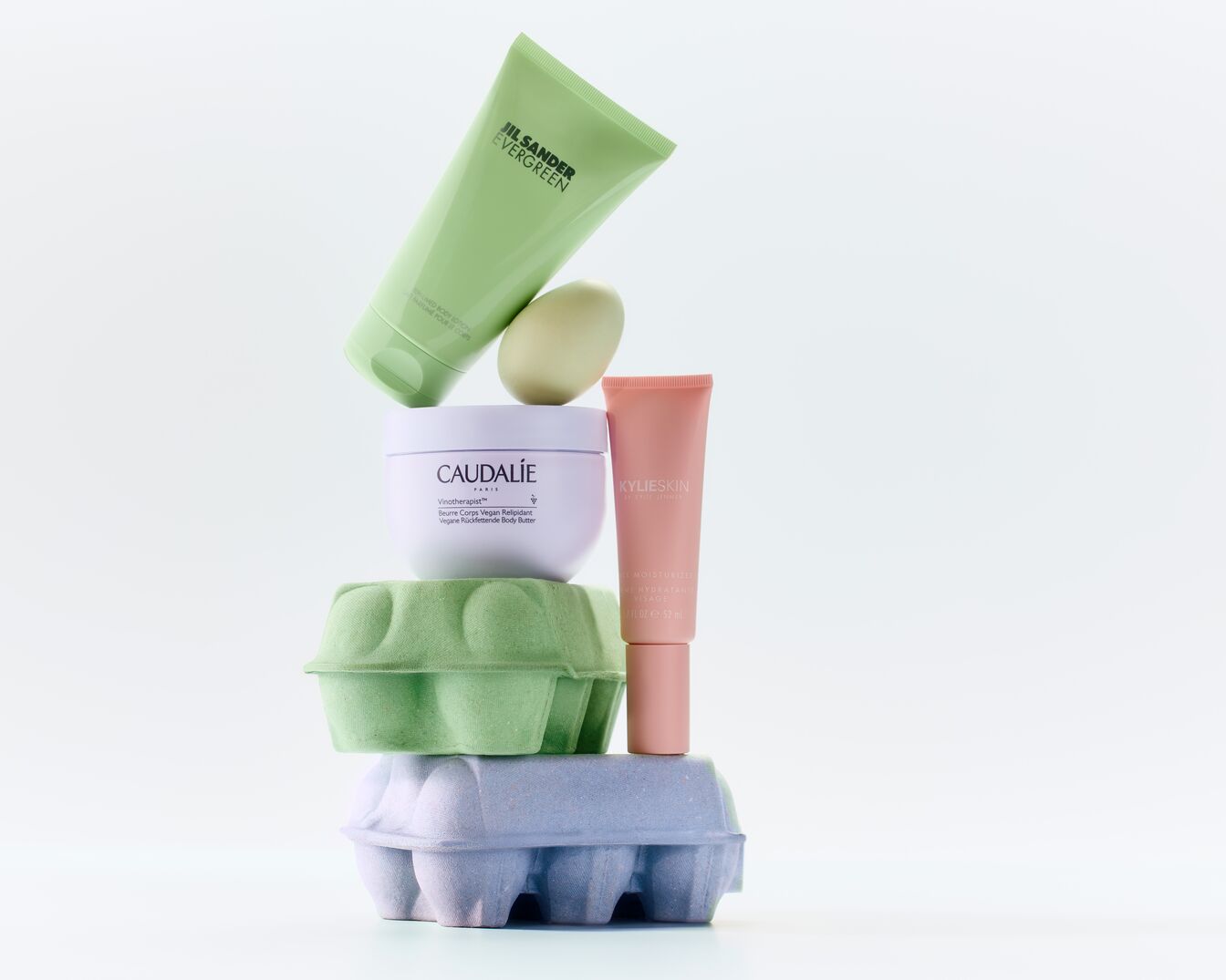 Ein Stapel von Schönheitsprodukten, darunter JIL SANDER EVERGREEN Lotion in einer hellgrünen Tube, Caudalie Körperbutter in einem weißen Tiegel und KYLIESKIN Feuchtigkeitscreme in einer rosa Tube, gestapelt auf pastellfarbenen Eierkartons.