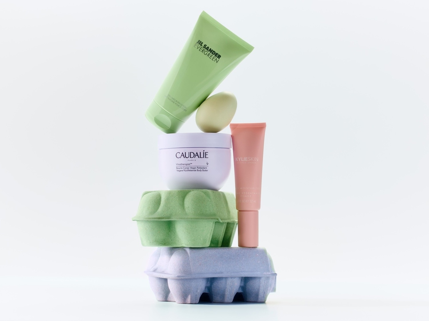 Ein Stapel von Schönheitsprodukten, darunter JIL SANDER EVERGREEN Lotion in grüner Tube, Caudalie Körperbutter in weißem Tiegel und Kylieskin Feuchtigkeitscreme in rosa Tube, gestapelt auf farbigen Eierkartons.