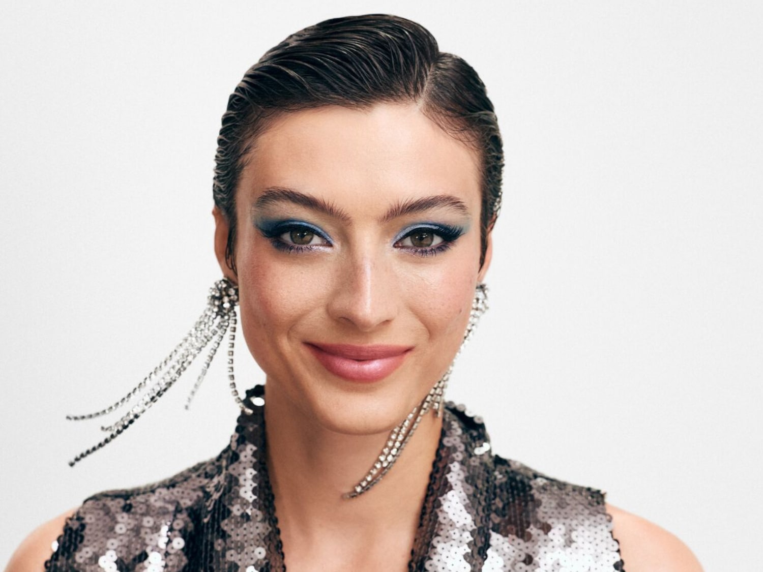 Portret kobiety z niebieskim makijażem oczu, błyszczącą sukienką i wiszącymi kolczykami, uśmiechającej się do kamery.