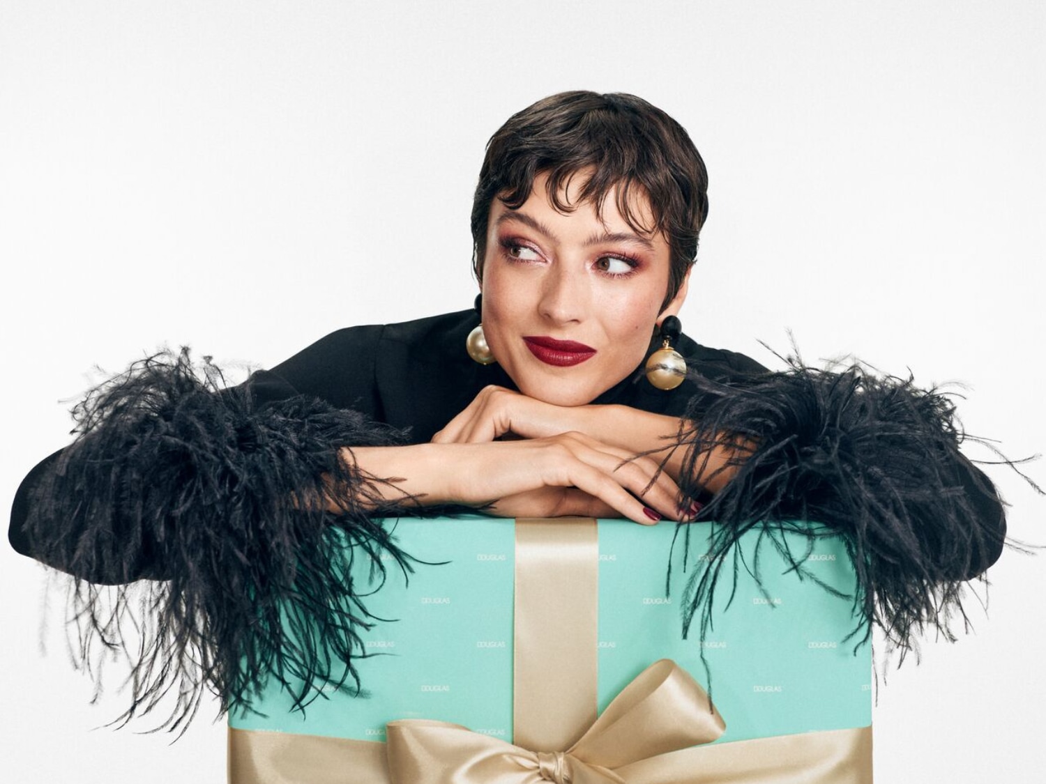 Kobieta z krótkimi, ciemnymi włosami i ciemną szminką opiera się na turkusowym pudełku prezentowym z kokardą w kolorze szampana, ubrana w czarną bluzkę z piórami.