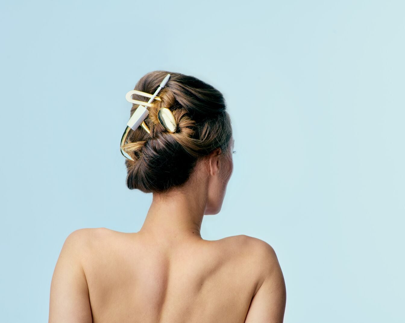 Kobieta z upiętymi brązowymi włosami, ozdobionymi złotą spinką, prezentuje elegancką fryzurę na tle błękitu.