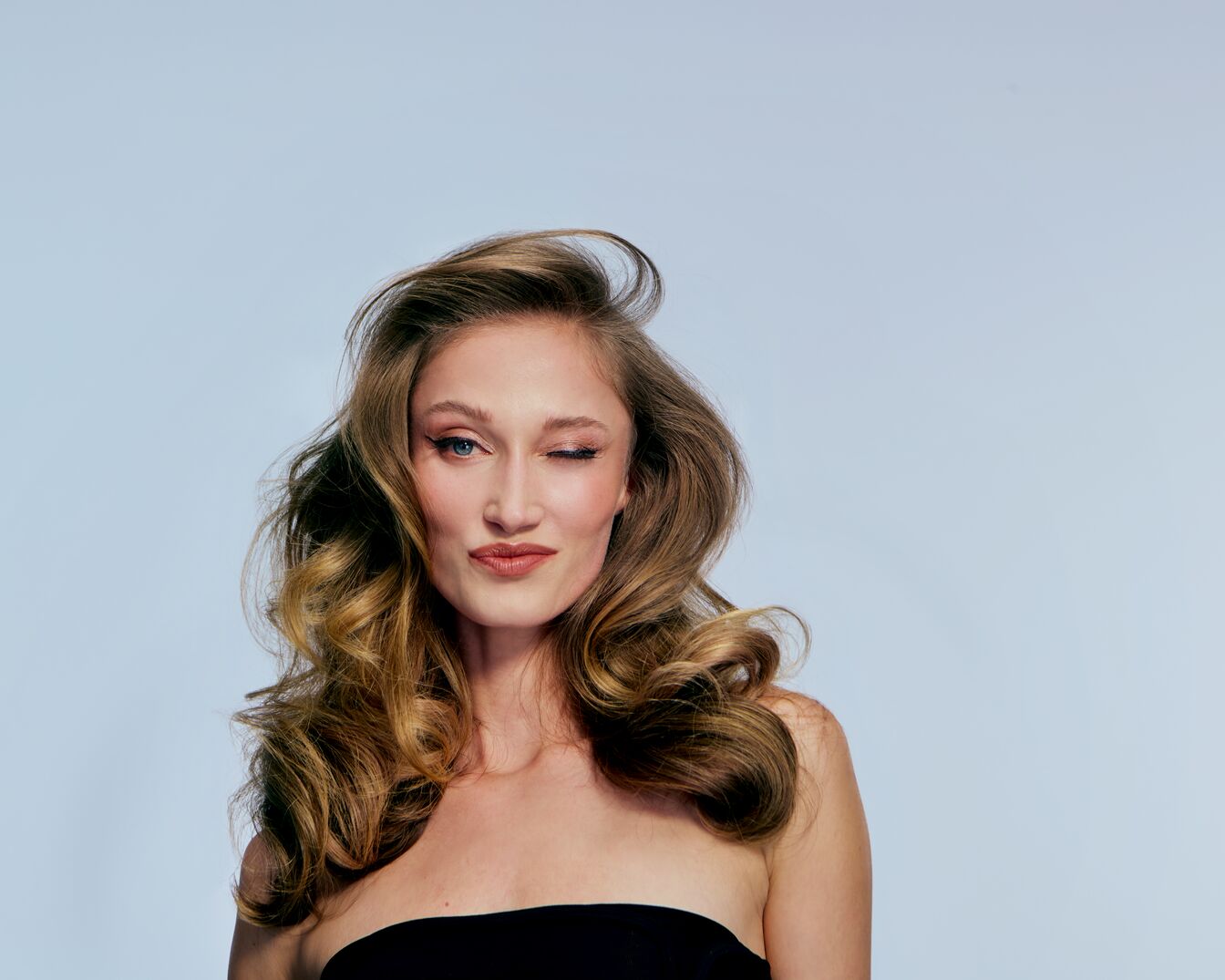 Portret kobiety z długimi, kręconymi włosami w odcieniach blondu i brązu, ubranej w czarny top bez ramiączek, która zalotnie puszcza oko.