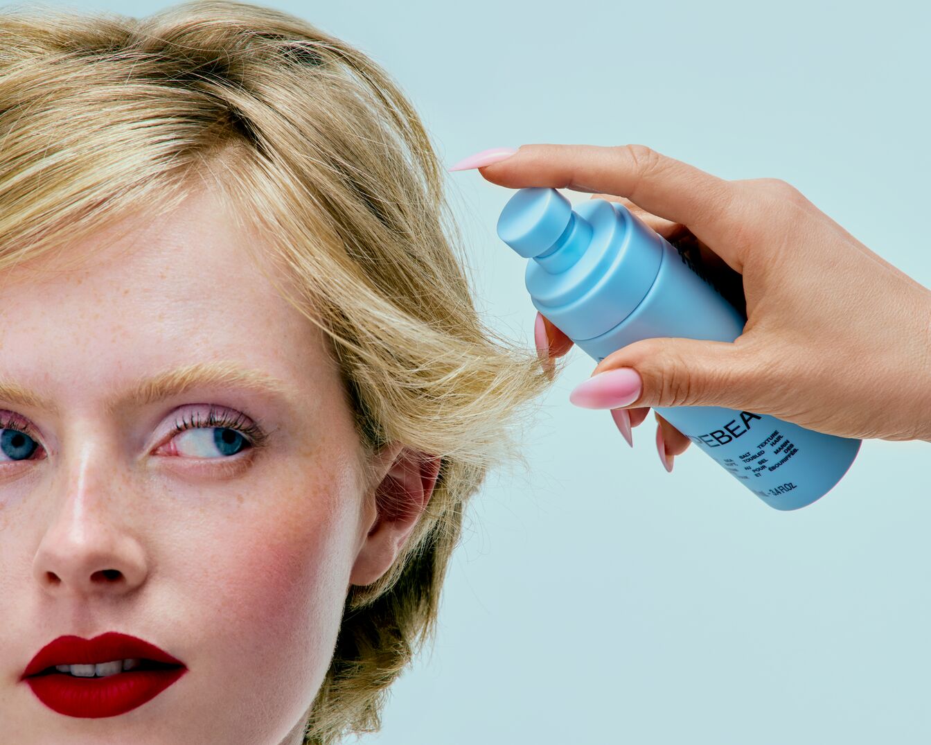 Kobieta z krótkimi blond włosami i czerwonymi ustami spryskuje włosy niebieską butelką produktu do stylizacji włosów, aby uzyskać teksturę i utrwalenie.