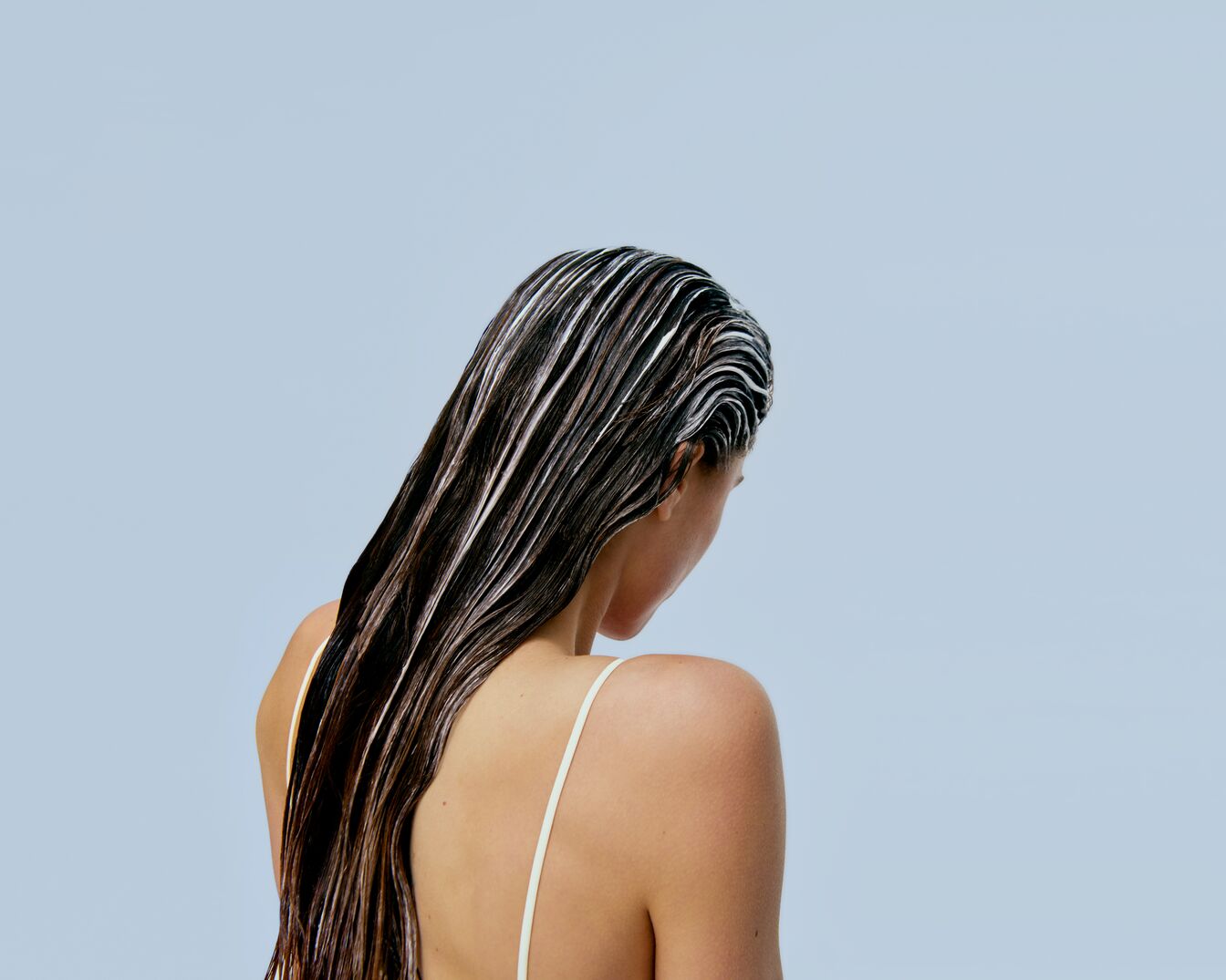 Kobieta w białym topie z nałożoną odżywką na ciemne, mokre włosy, stojąca tyłem na jasnoniebieskim tle.