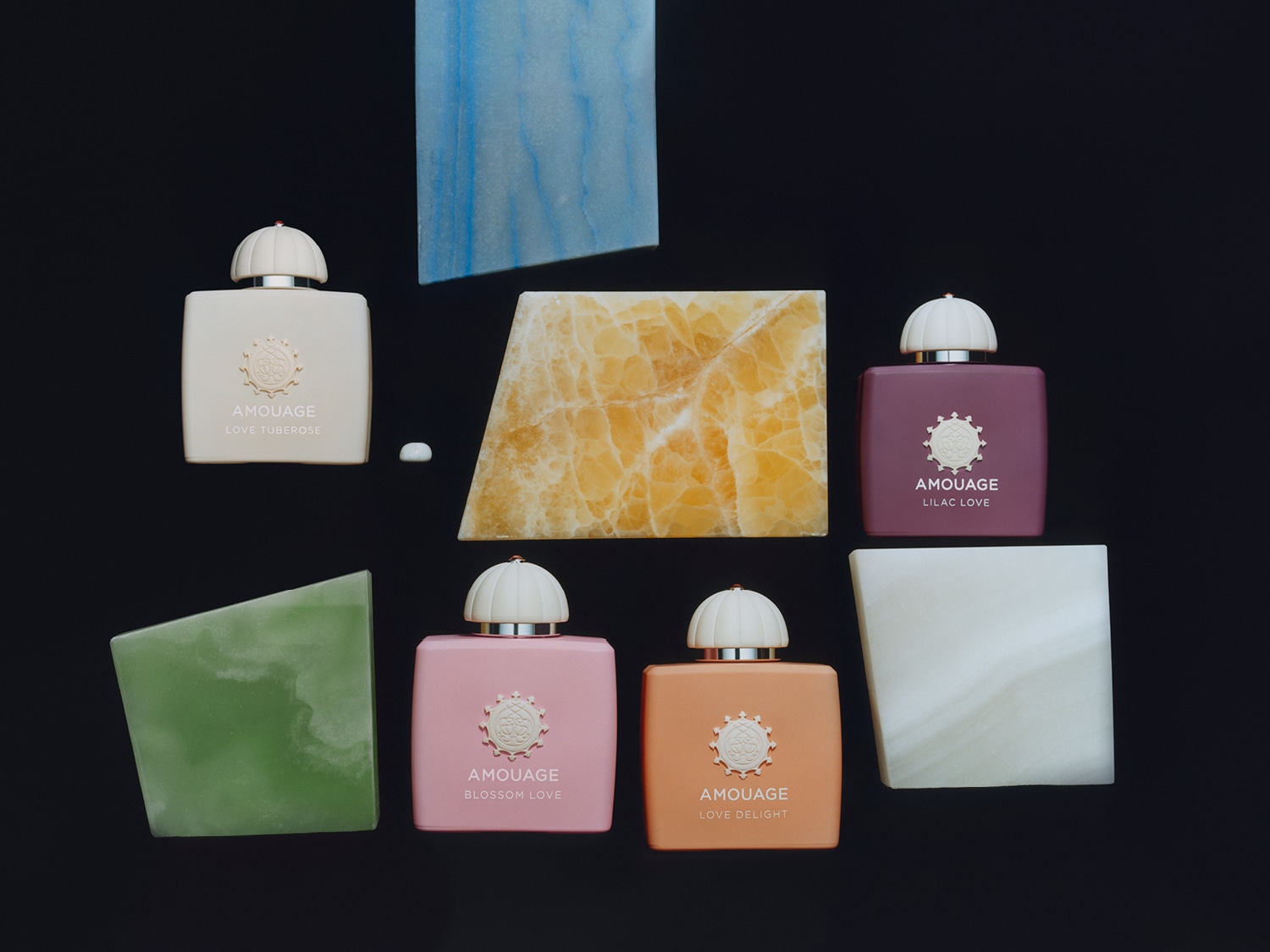 Quadratische Parfümflakons von Amouage in Weiß, Lila, Rosa und Orange, jeweils mit dem Namen des Duftes, umgeben von farbigen Marmorstücken auf schwarzem Hintergrund.